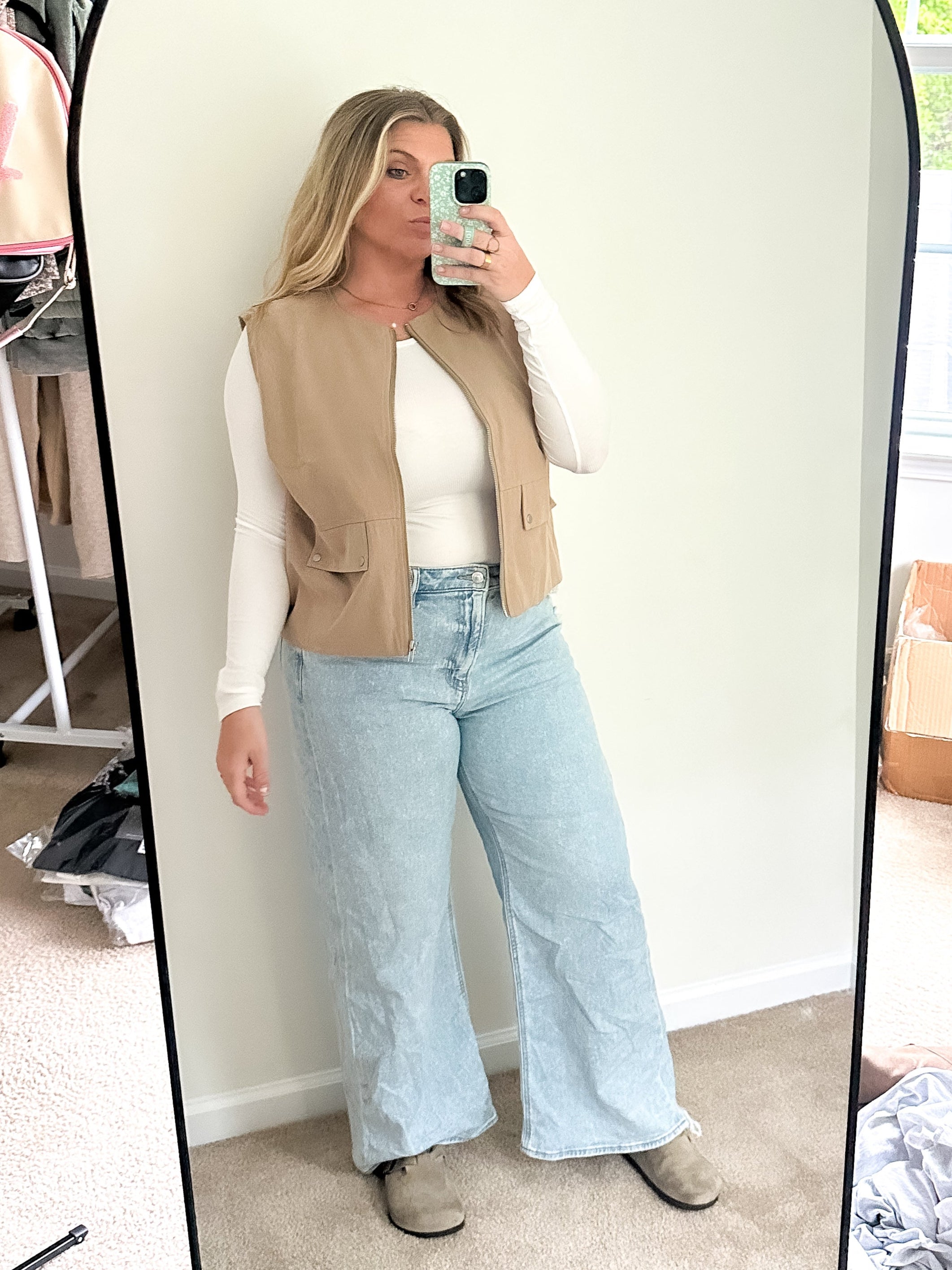 Person taking a mirror selfie wearing a beige vest, white shirt, and light blue jeans in a room with a window and boxes.