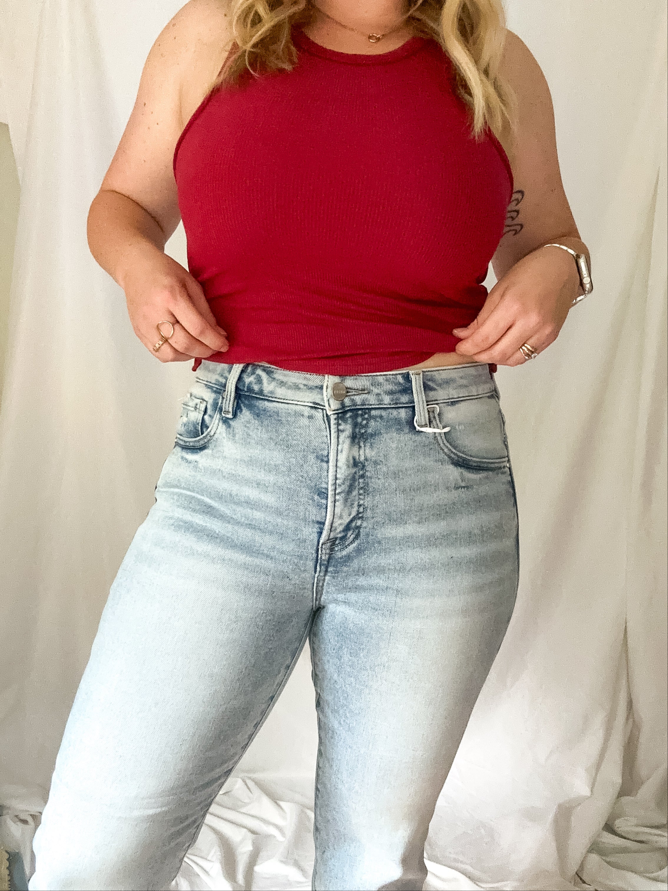 Person wearing a red tank top and light blue jeans against a plain background