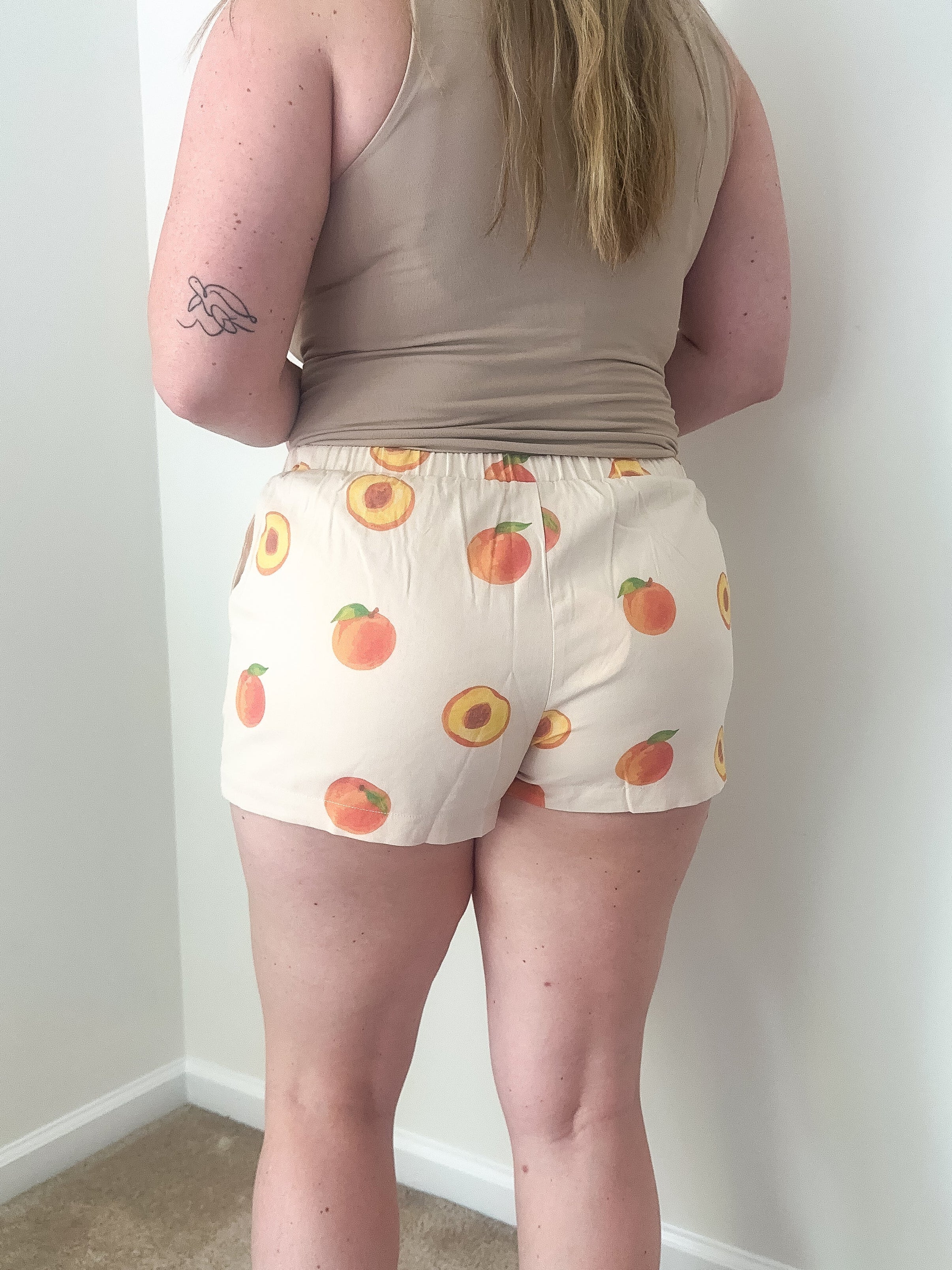 Person wearing a beige tank top and shorts with peach pattern against a white wall.