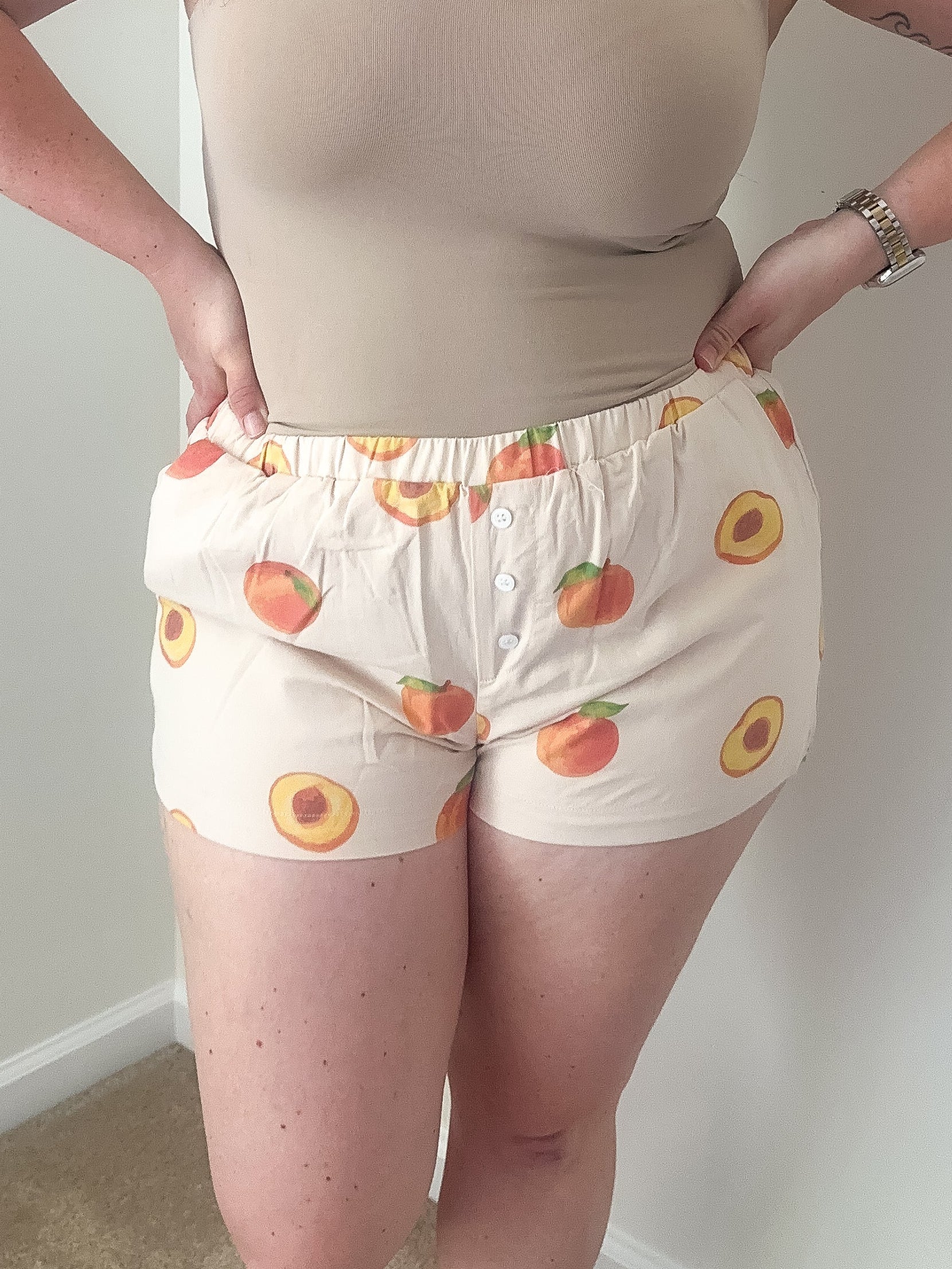 Woman wearing a beige tank top and patterned shorts against a plain wall.