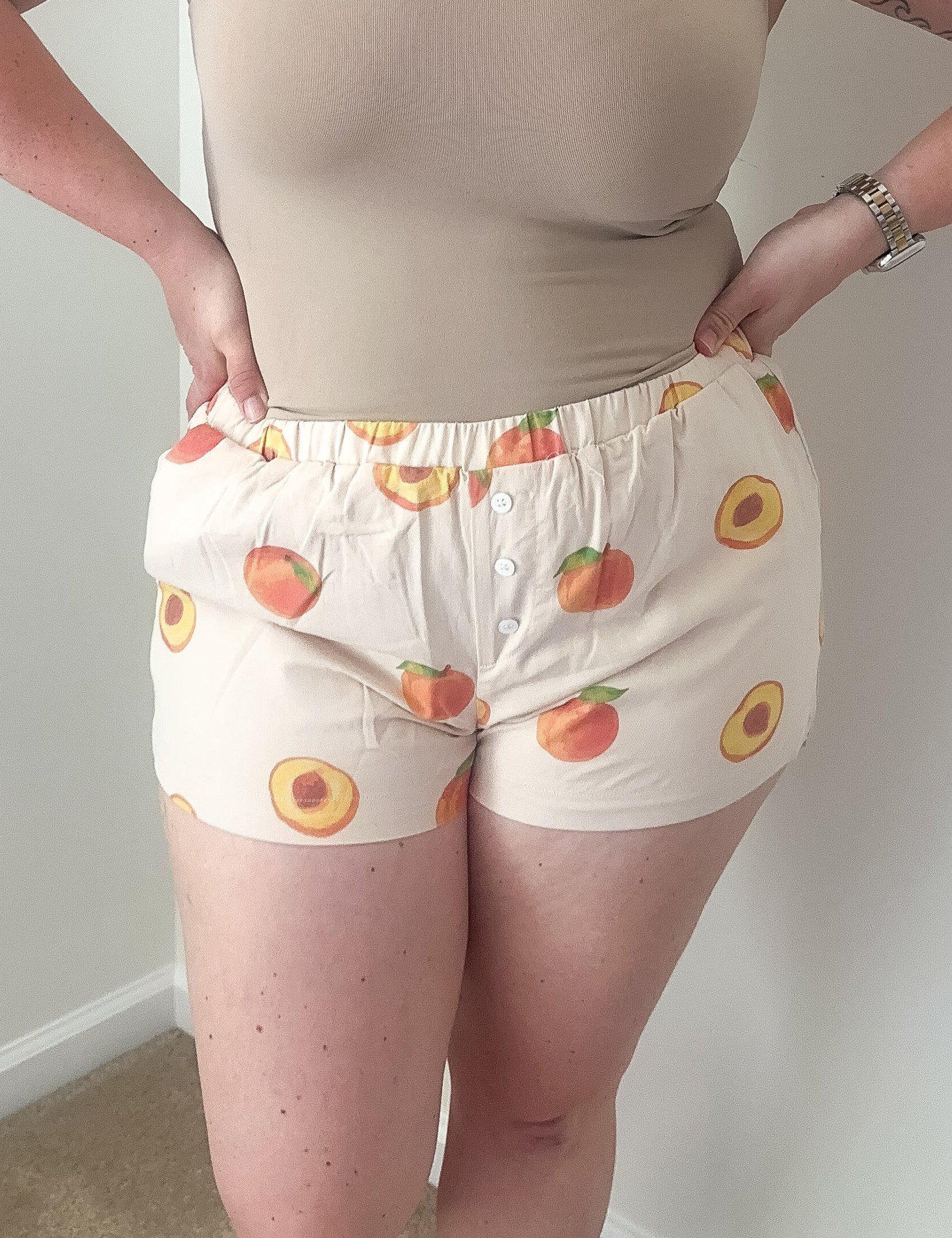 Woman wearing a beige tank top and patterned shorts against a plain wall.