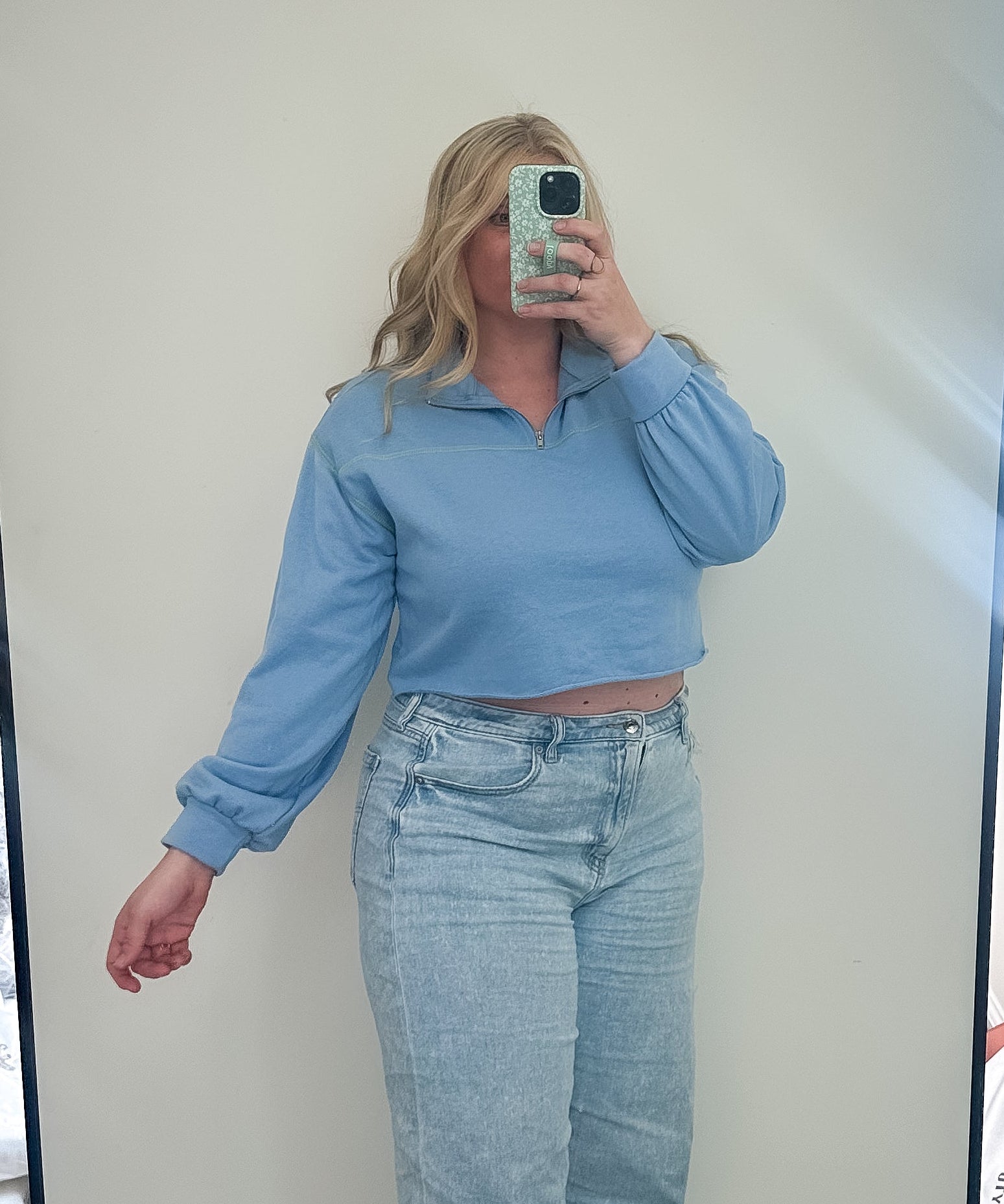 Person taking a mirror selfie wearing a blue top and light blue jeans in a room with a large mirror.