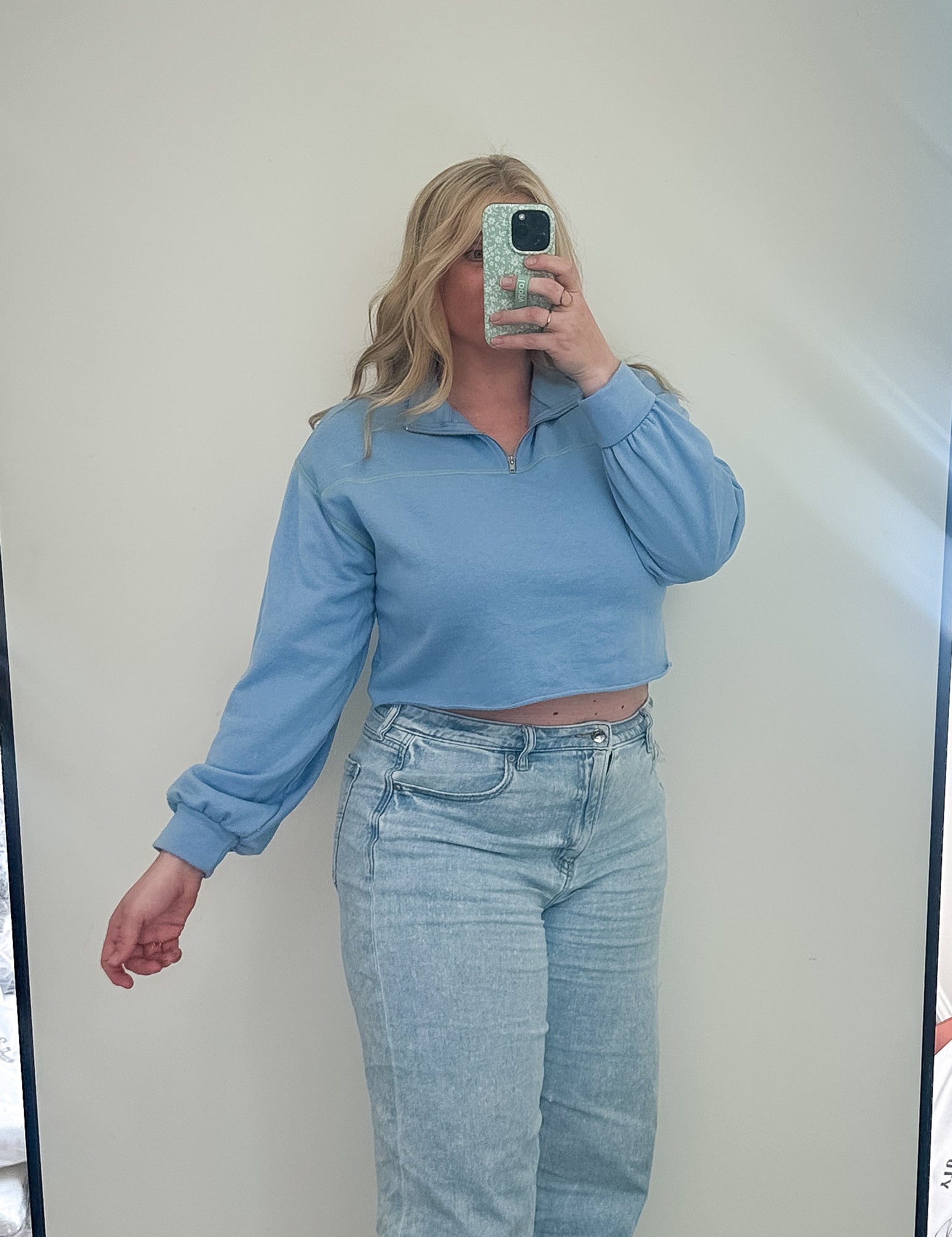 Person taking a mirror selfie wearing a blue top and light blue jeans in a room with a large mirror.