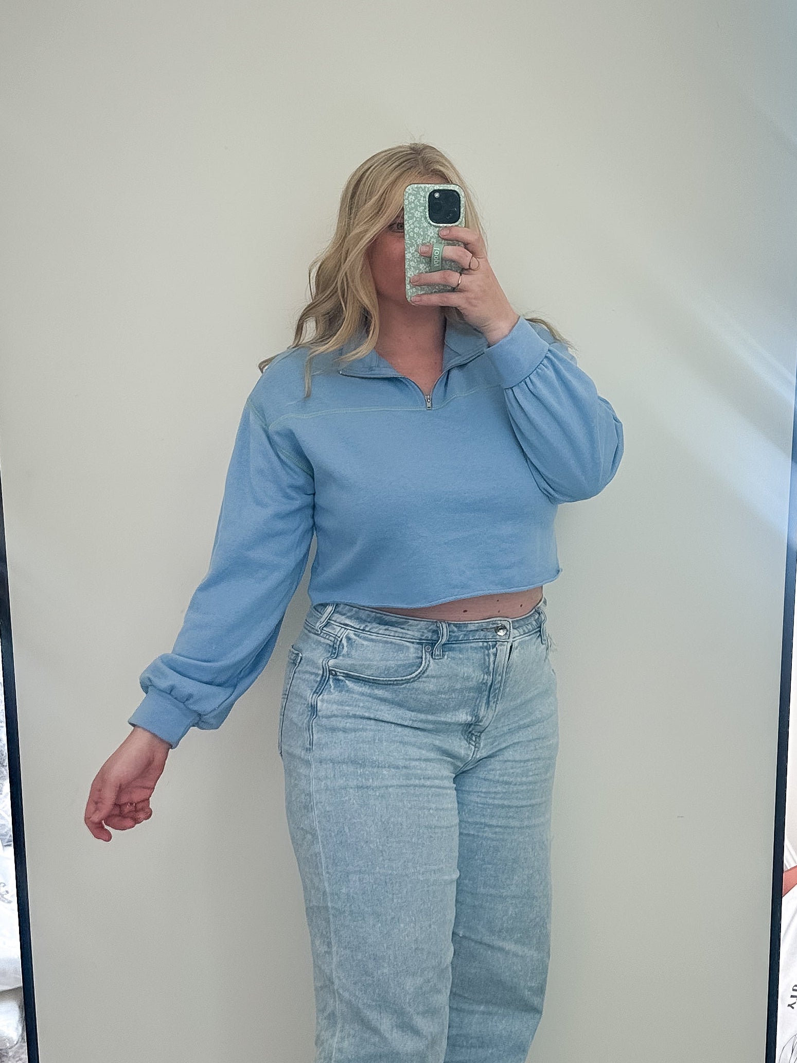 Person taking a mirror selfie wearing a blue top and light blue jeans in a room with a large mirror.