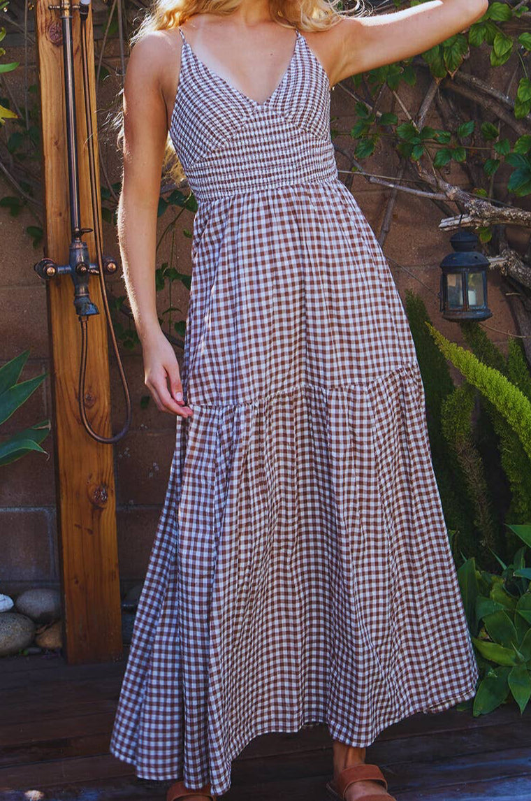 Woman wearing a checkered dress standing outdoors with plants and a wooden structure in the background