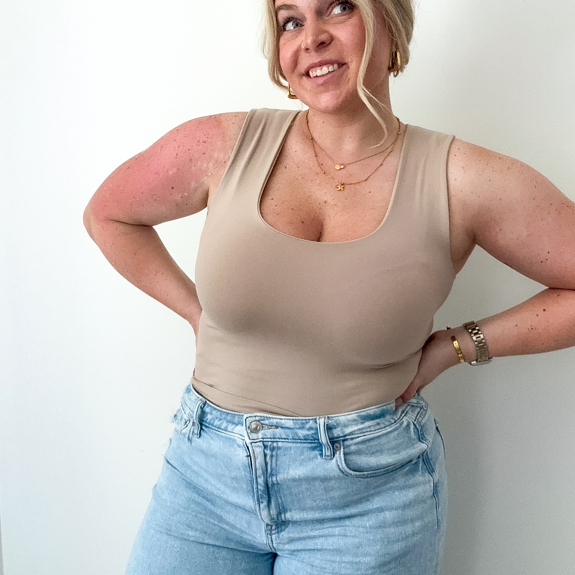 Woman wearing a beige tank top and light blue jeans against a white wall