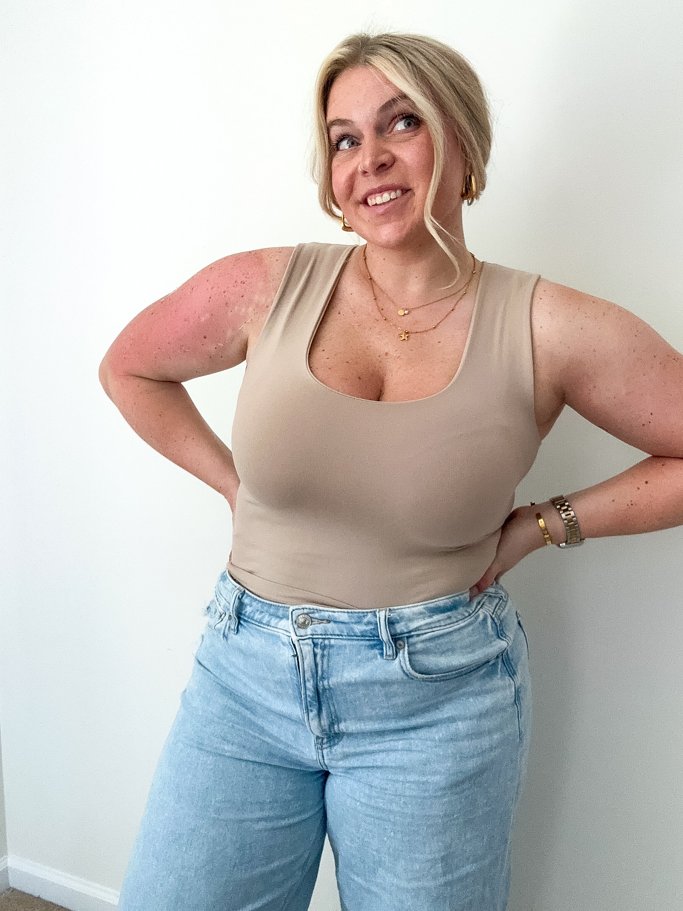 Woman wearing a beige tank top and light blue jeans against a white wall