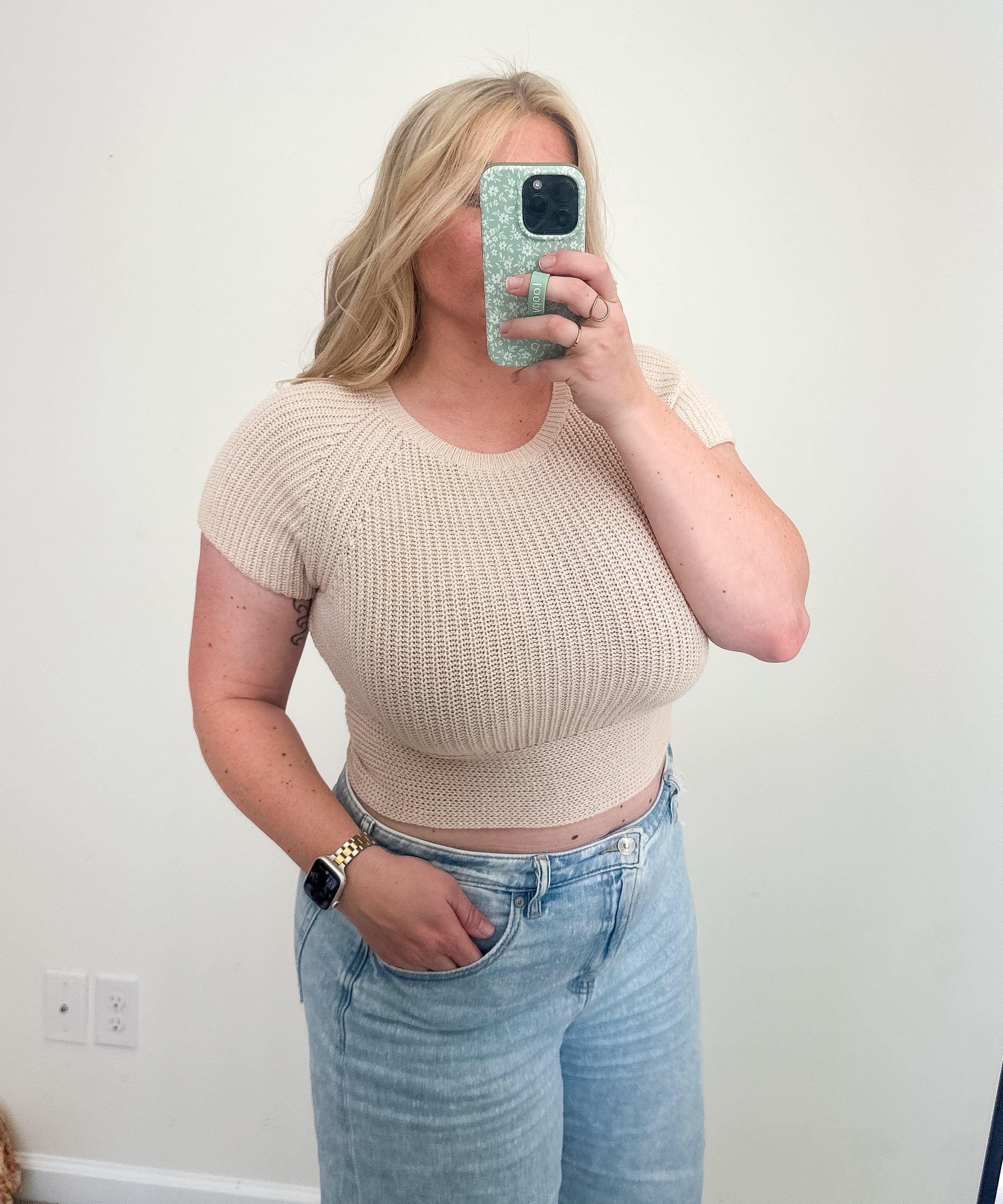 Person wearing a beige cropped top and light blue jeans taking a mirror selfie.