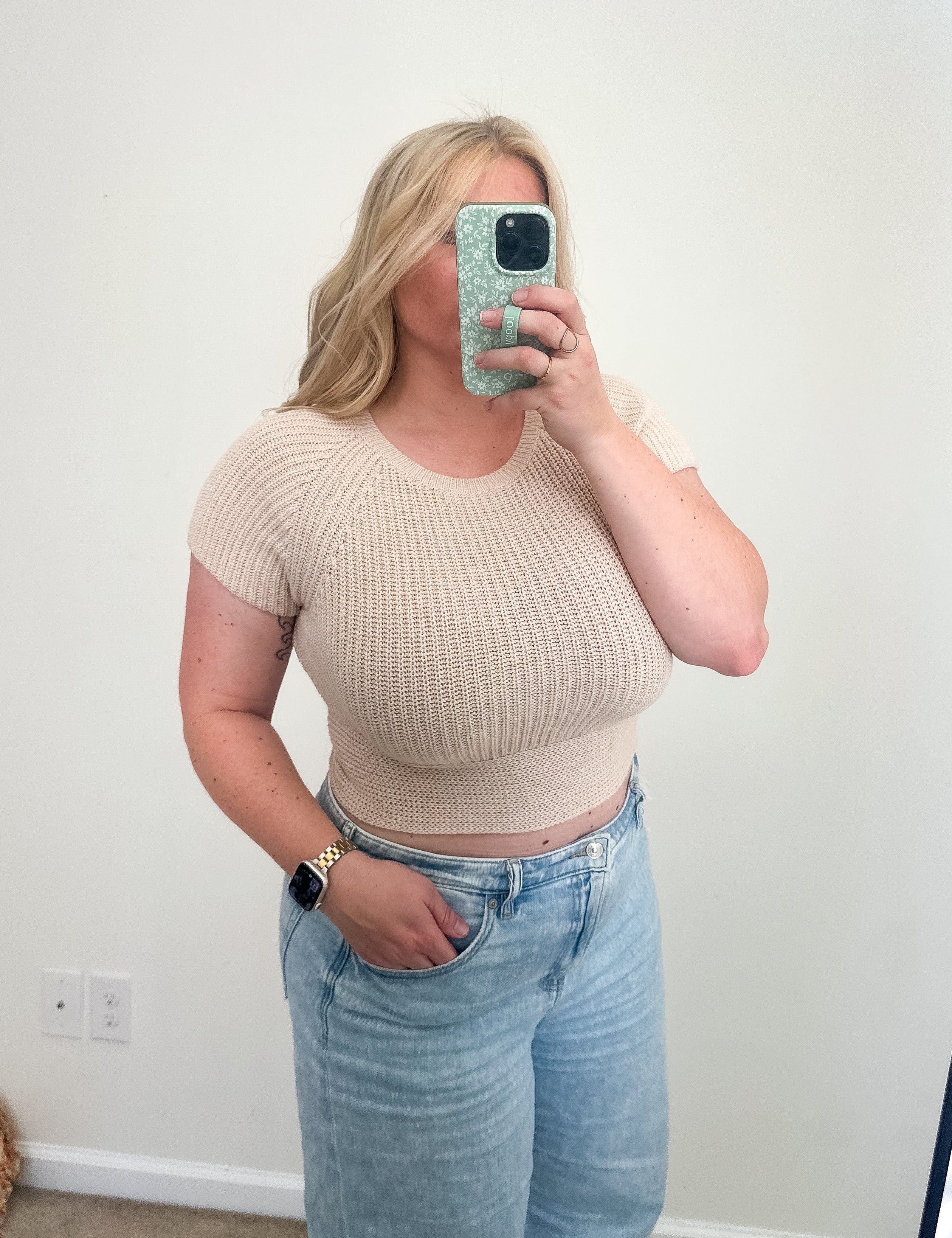 Person wearing a beige cropped top and light blue jeans taking a mirror selfie.