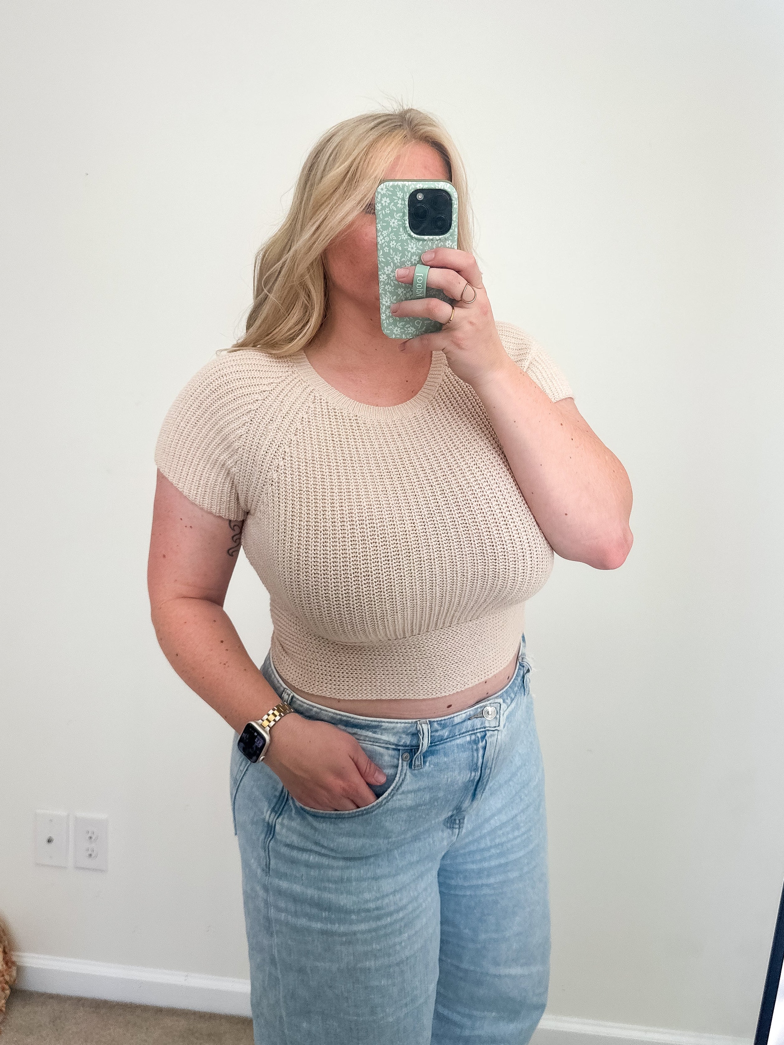 Person wearing a beige cropped top and light blue jeans taking a mirror selfie.