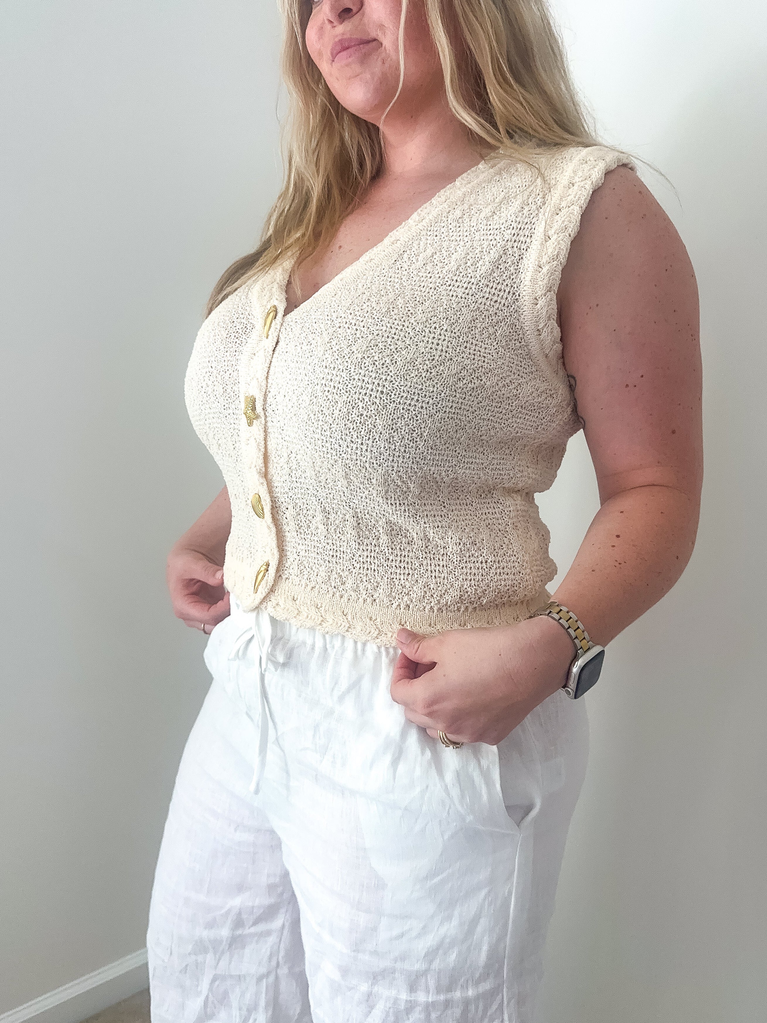 Woman wearing a cream-colored sleeveless knitted top and white pants against a plain background