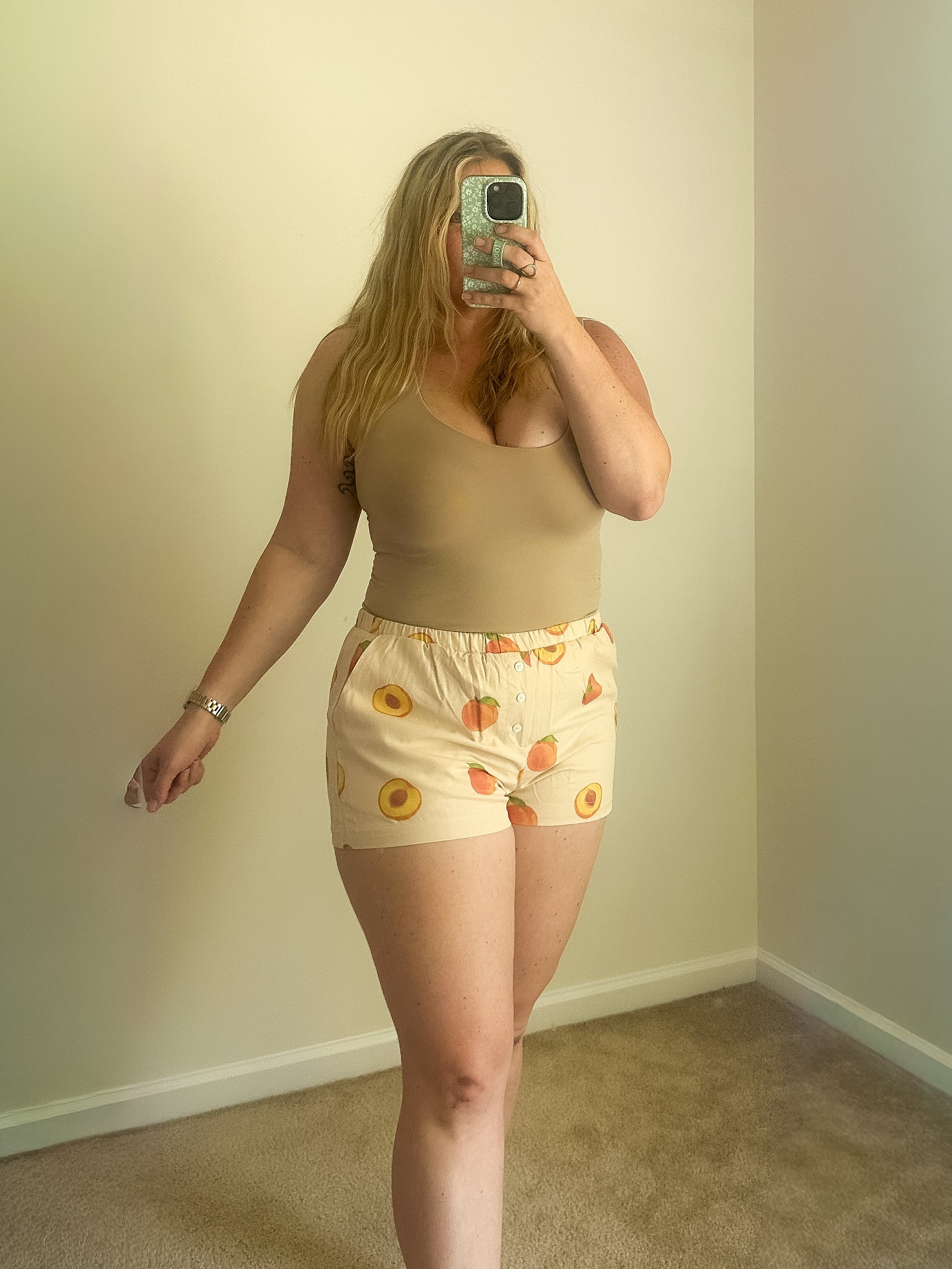 Person taking a mirror selfie wearing a beige top and patterned shorts in a room with plain walls.