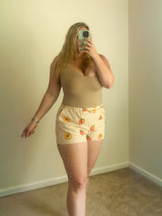 Person taking a mirror selfie wearing a beige top and patterned shorts in a room with plain walls.