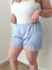 Person wearing a white top and light blue shorts with lace trim against a plain wall.