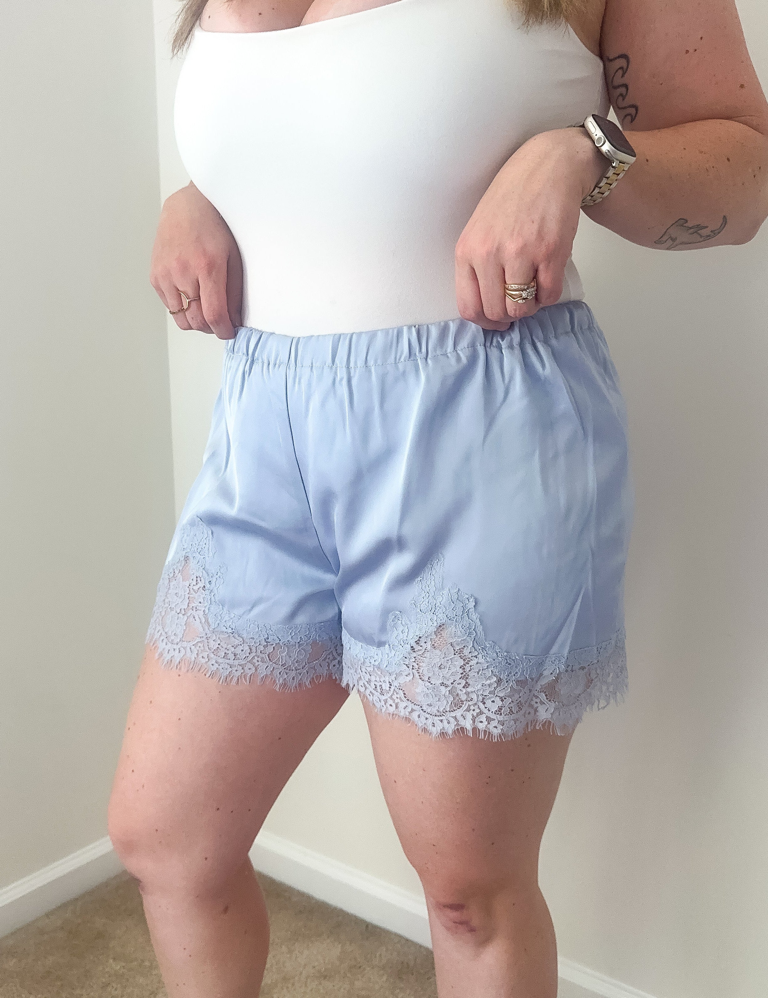 Person wearing a white top and light blue shorts with lace trim against a plain wall.