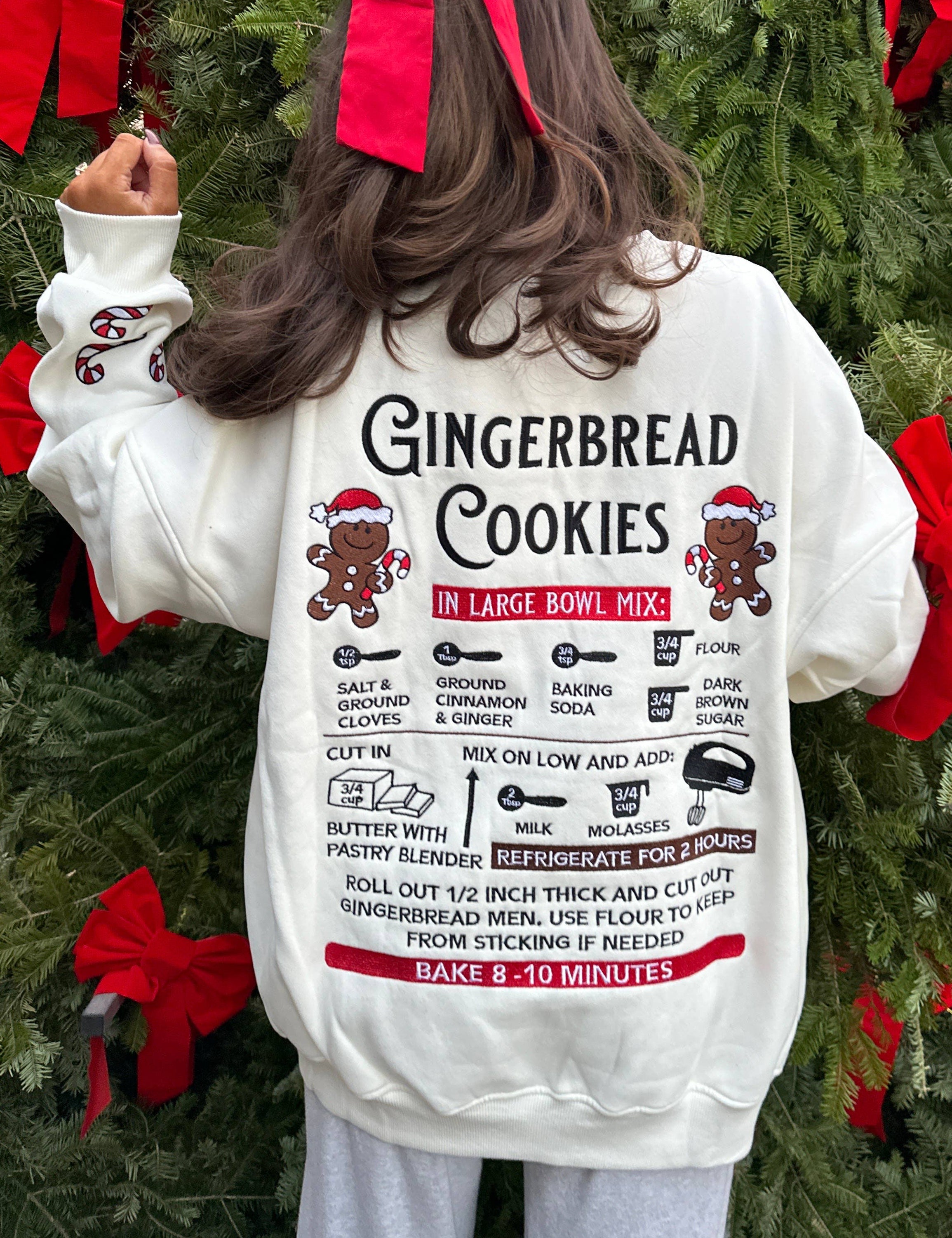 Person wearing a white sweatshirt with gingerbread cookies recipe design in front of a decorated Christmas tree.