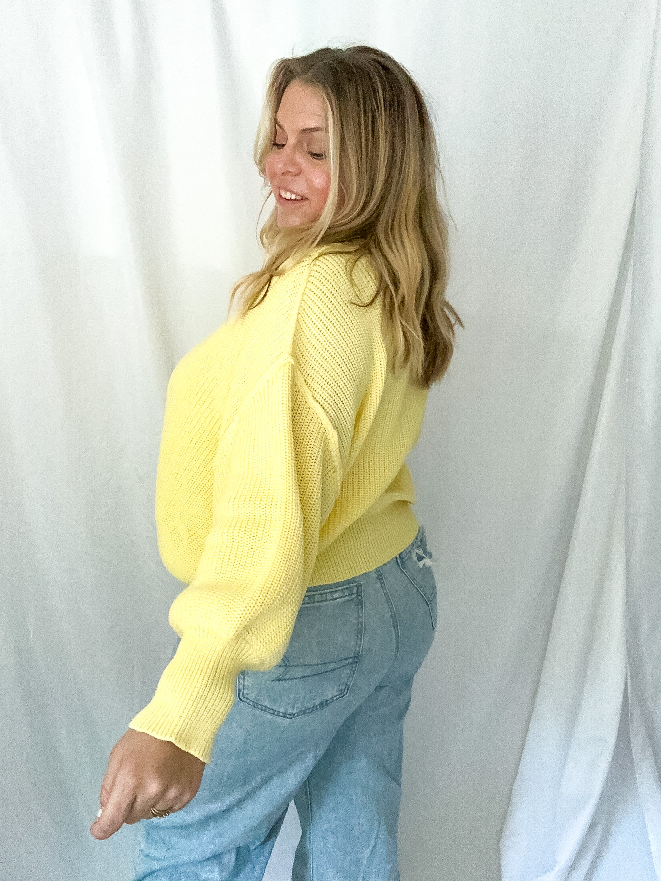 Woman wearing a yellow sweater and blue jeans against a white curtain background