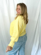 Woman wearing a yellow sweater and blue jeans against a white curtain background