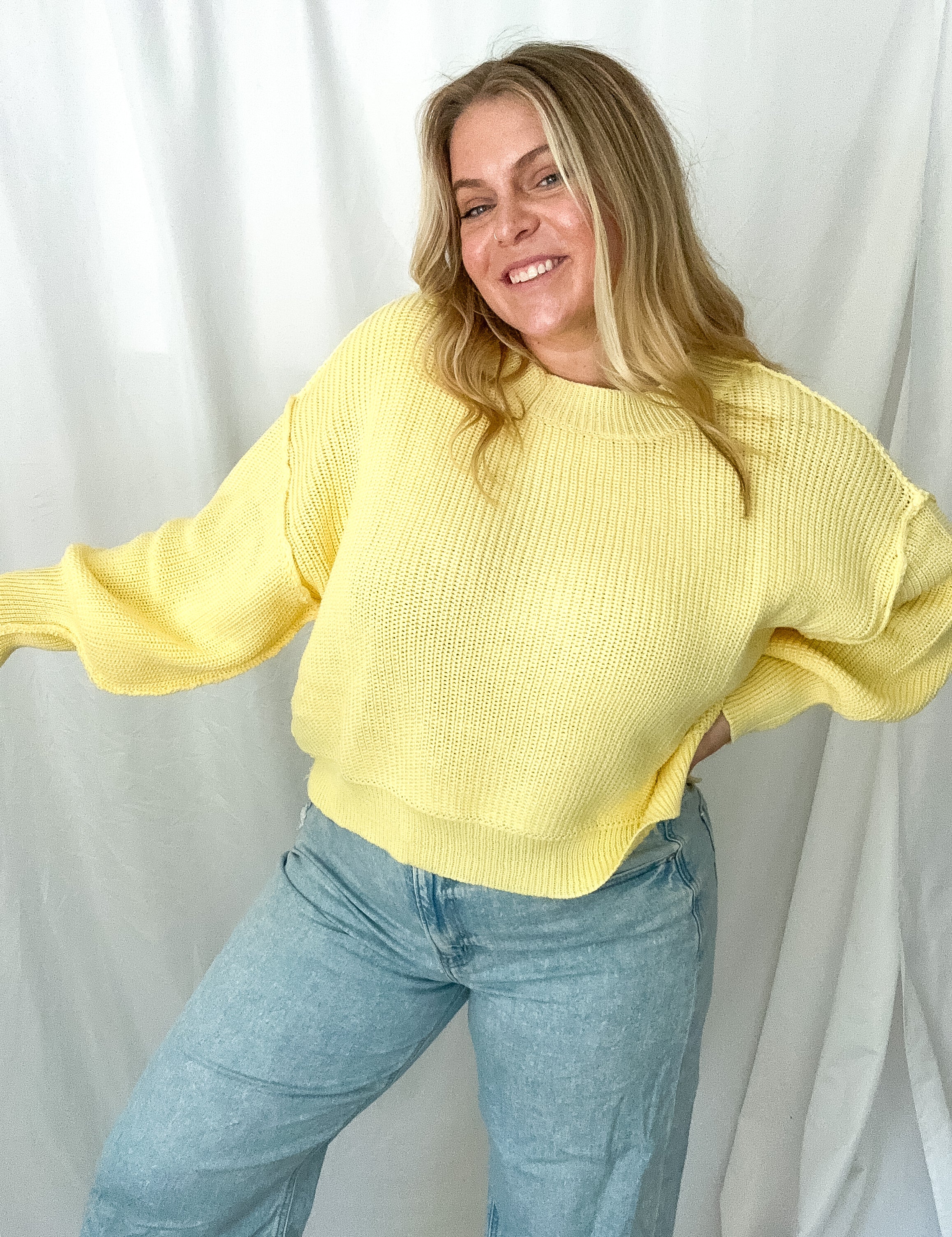 Person wearing a yellow sweater and blue jeans against a white curtain background