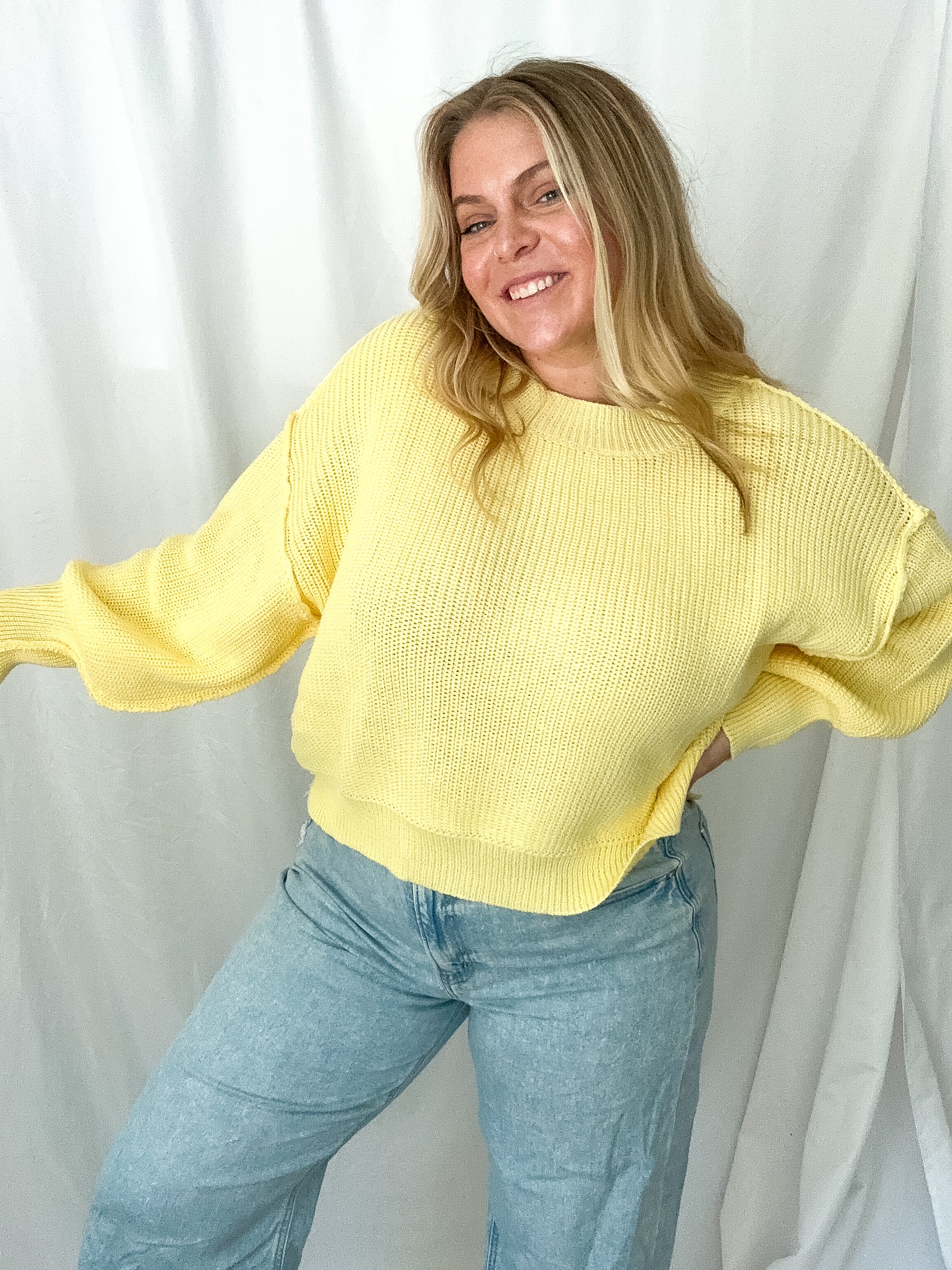 Person wearing a yellow sweater and blue jeans against a white curtain background
