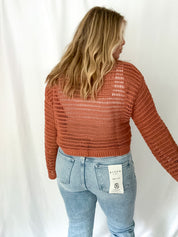 Person wearing a rust-colored knit sweater and light blue jeans with a white background