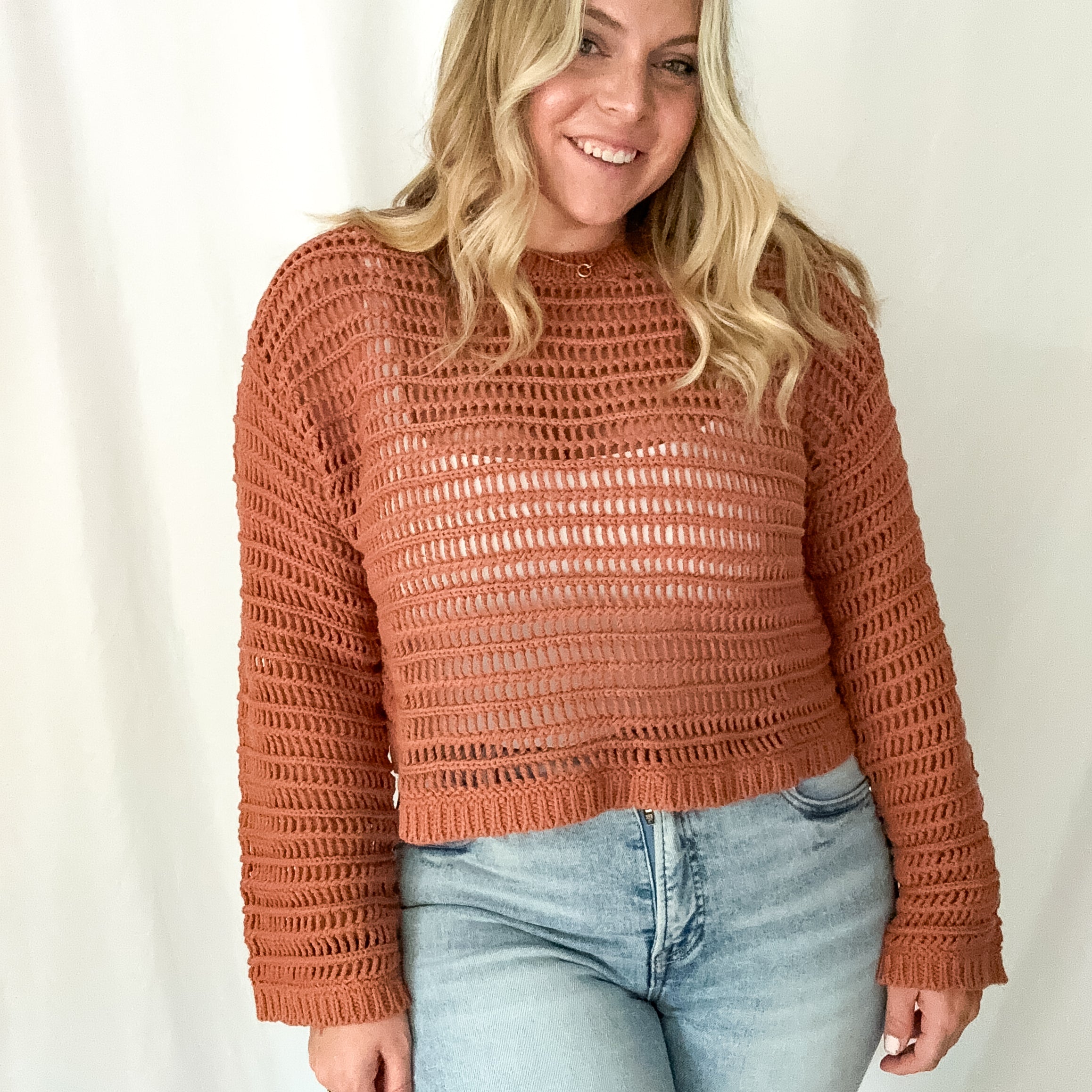Woman wearing a brown crochet sweater and light blue jeans against a white background