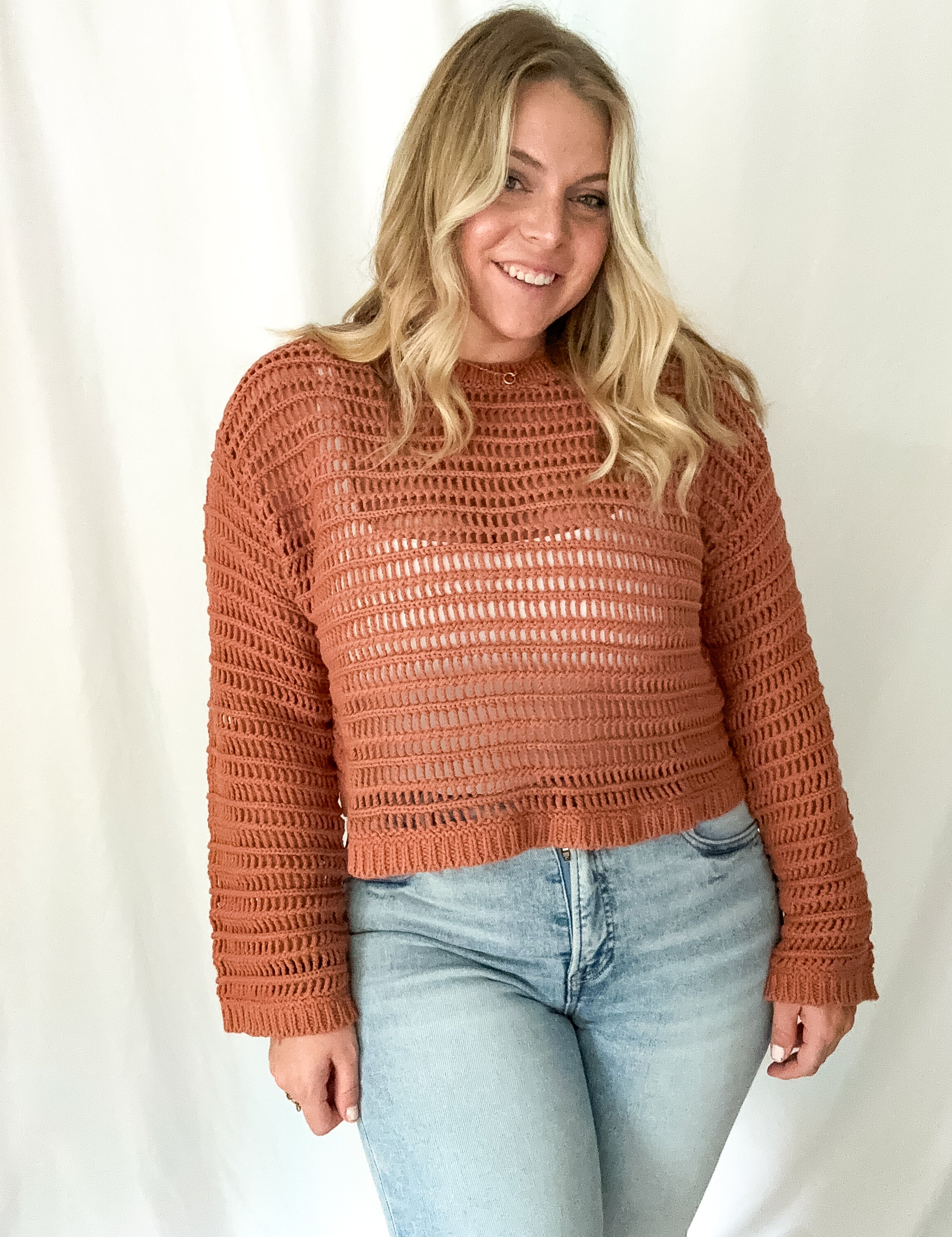 Woman wearing a brown crochet sweater and light blue jeans against a white background