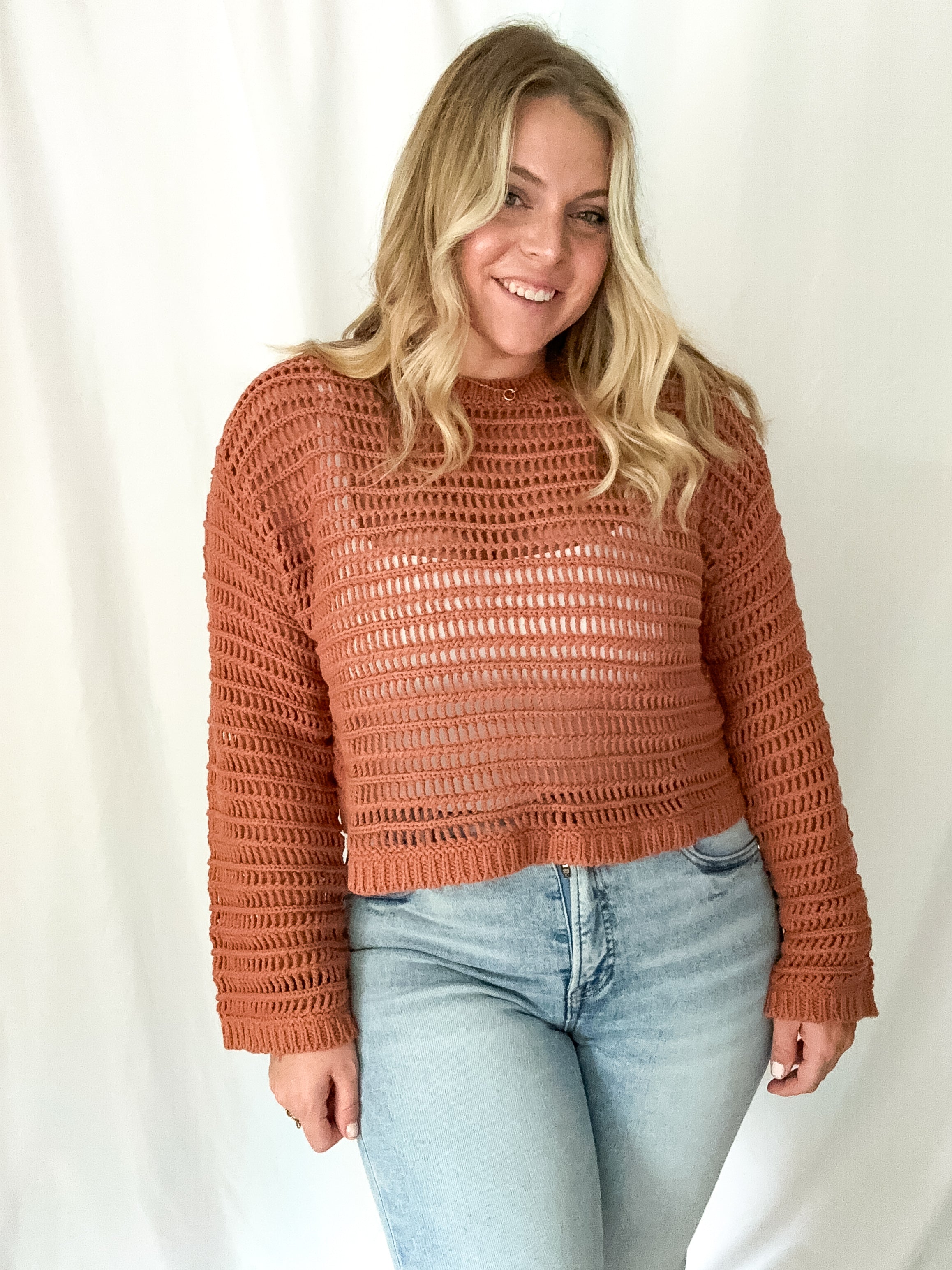 Woman wearing a brown crochet sweater and light blue jeans against a white background