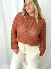Person wearing a brown knitted sweater and white pants against a plain background