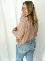 Woman wearing a beige cable knit sweater and light blue jeans against a white background