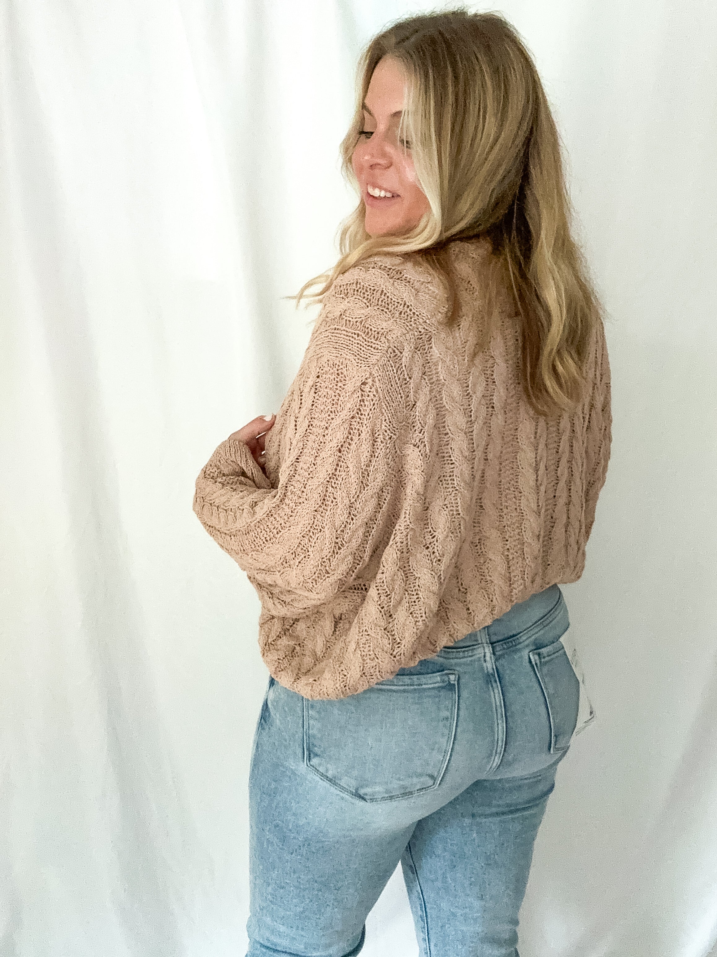 Woman wearing a beige cable knit sweater and light blue jeans against a white background