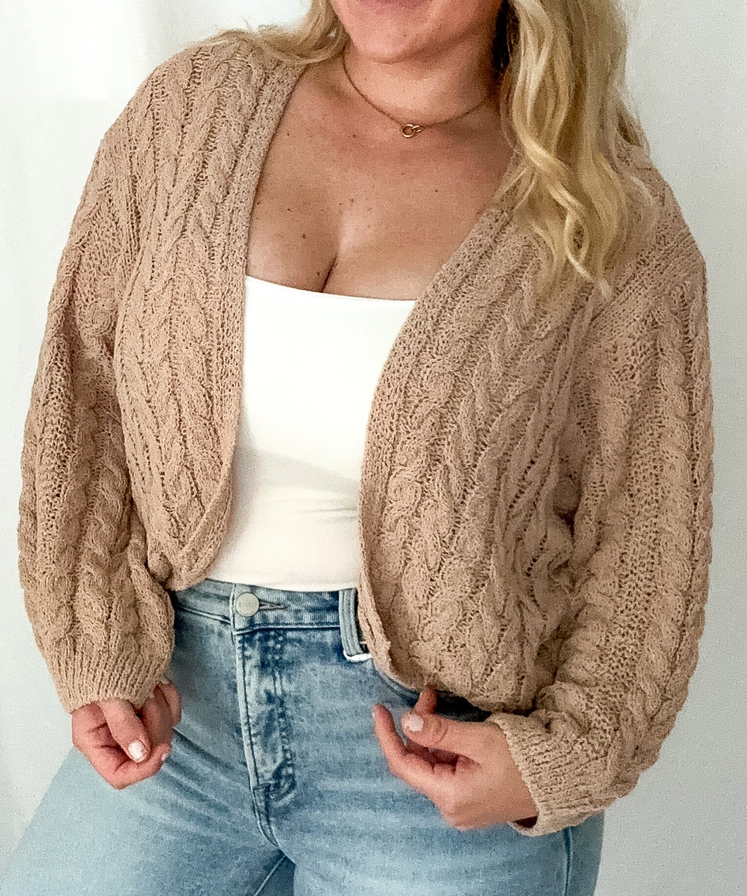 Woman wearing a beige cable knit cardigan over a white top and light blue jeans on a plain background
