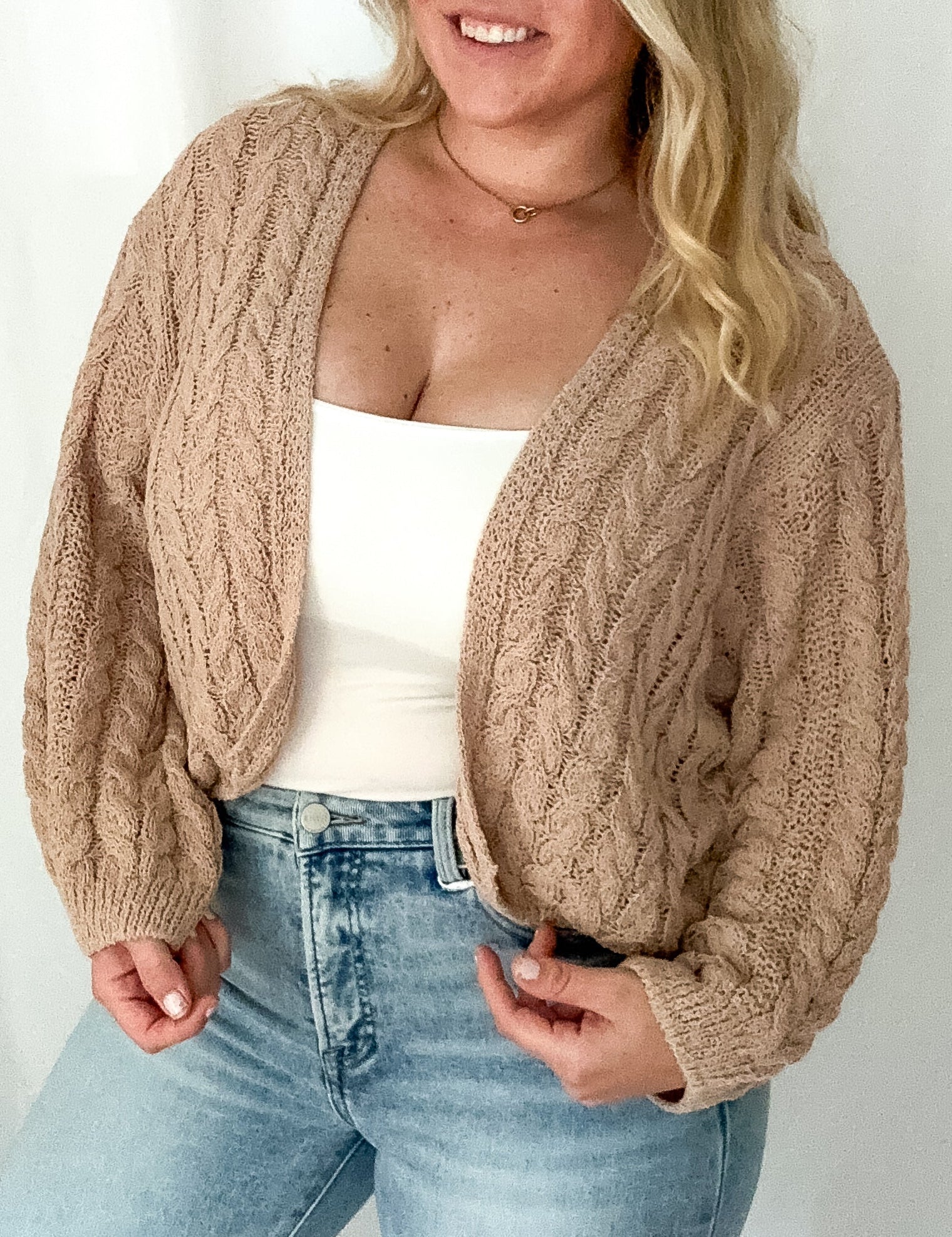 Woman wearing a beige cable knit cardigan over a white top and light blue jeans on a plain background