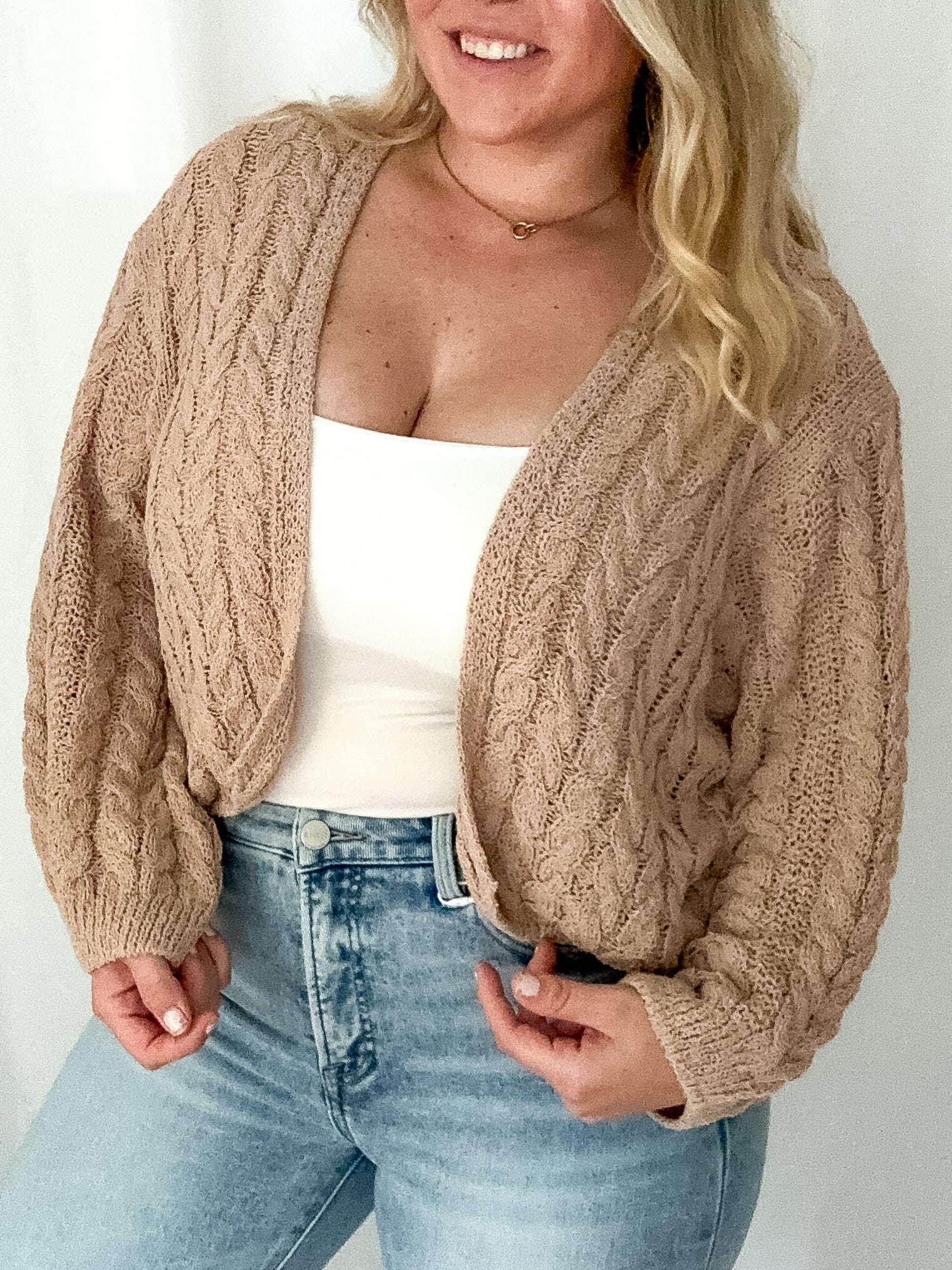 Woman wearing a beige cable knit cardigan over a white top and light blue jeans on a plain background