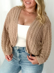 Woman wearing a beige cable knit cardigan over a white top and light blue jeans on a plain background