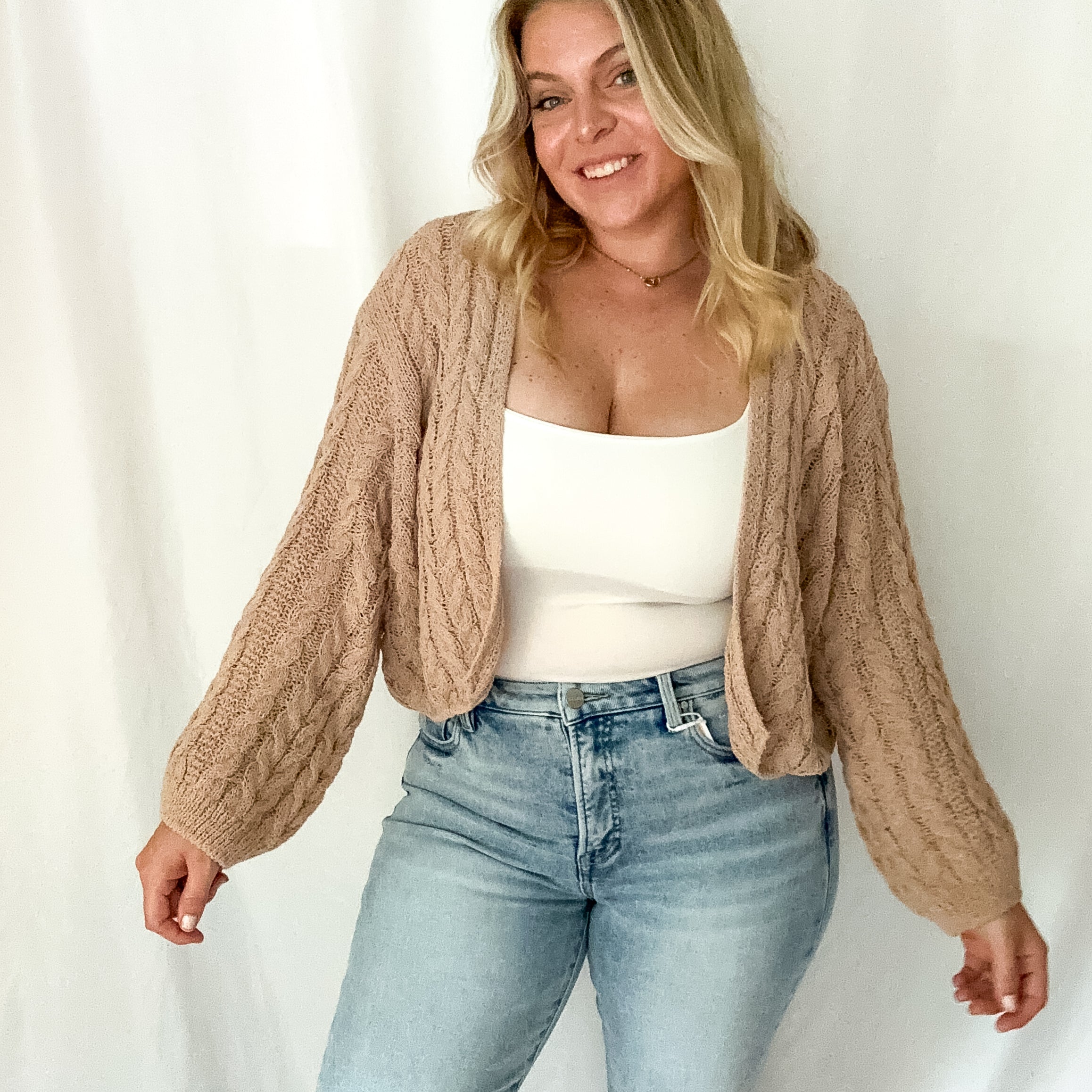 Woman wearing a beige cable knit cardigan, white top, and light blue jeans against a white background