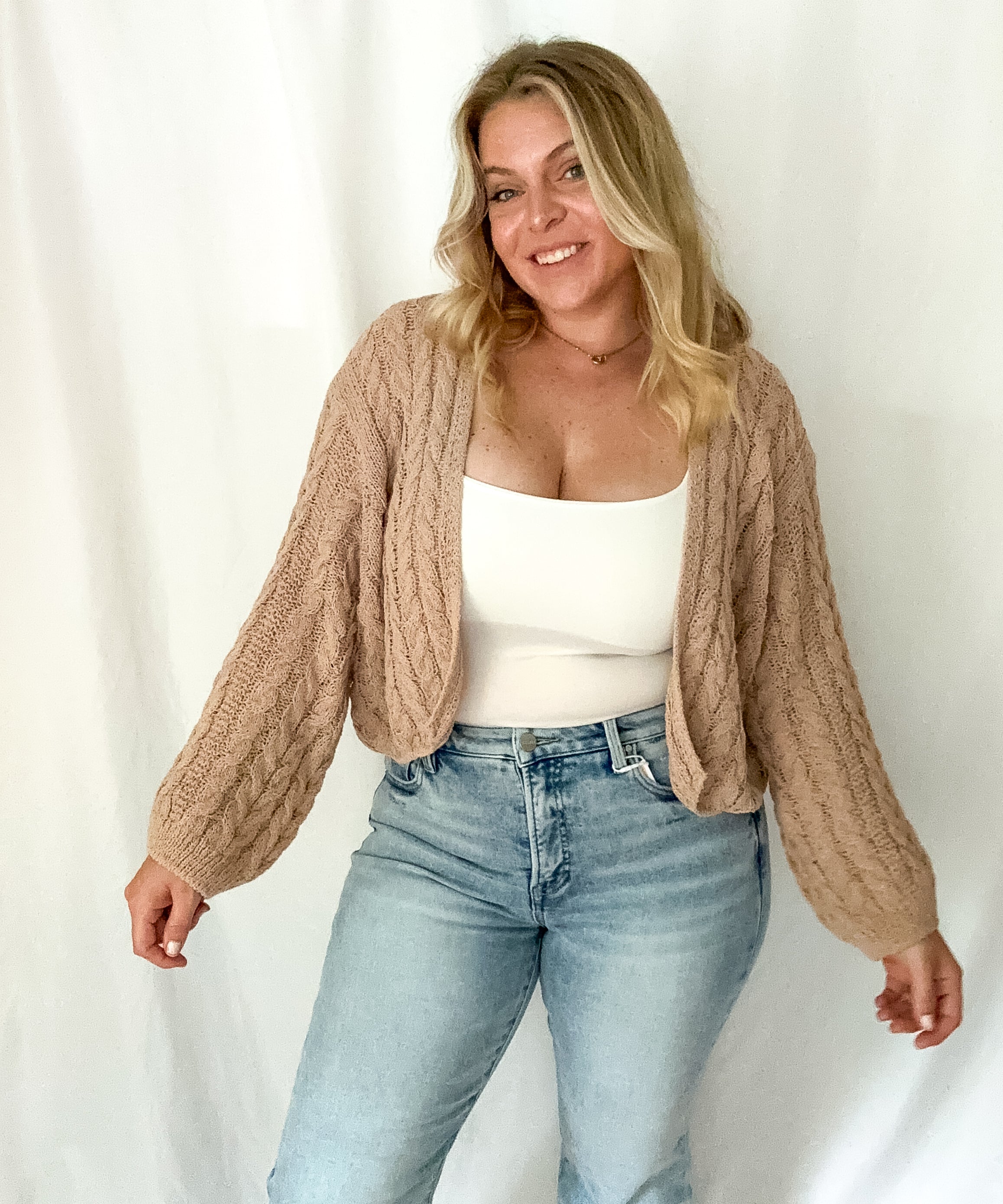 Woman wearing a beige cable knit cardigan, white top, and light blue jeans against a white background