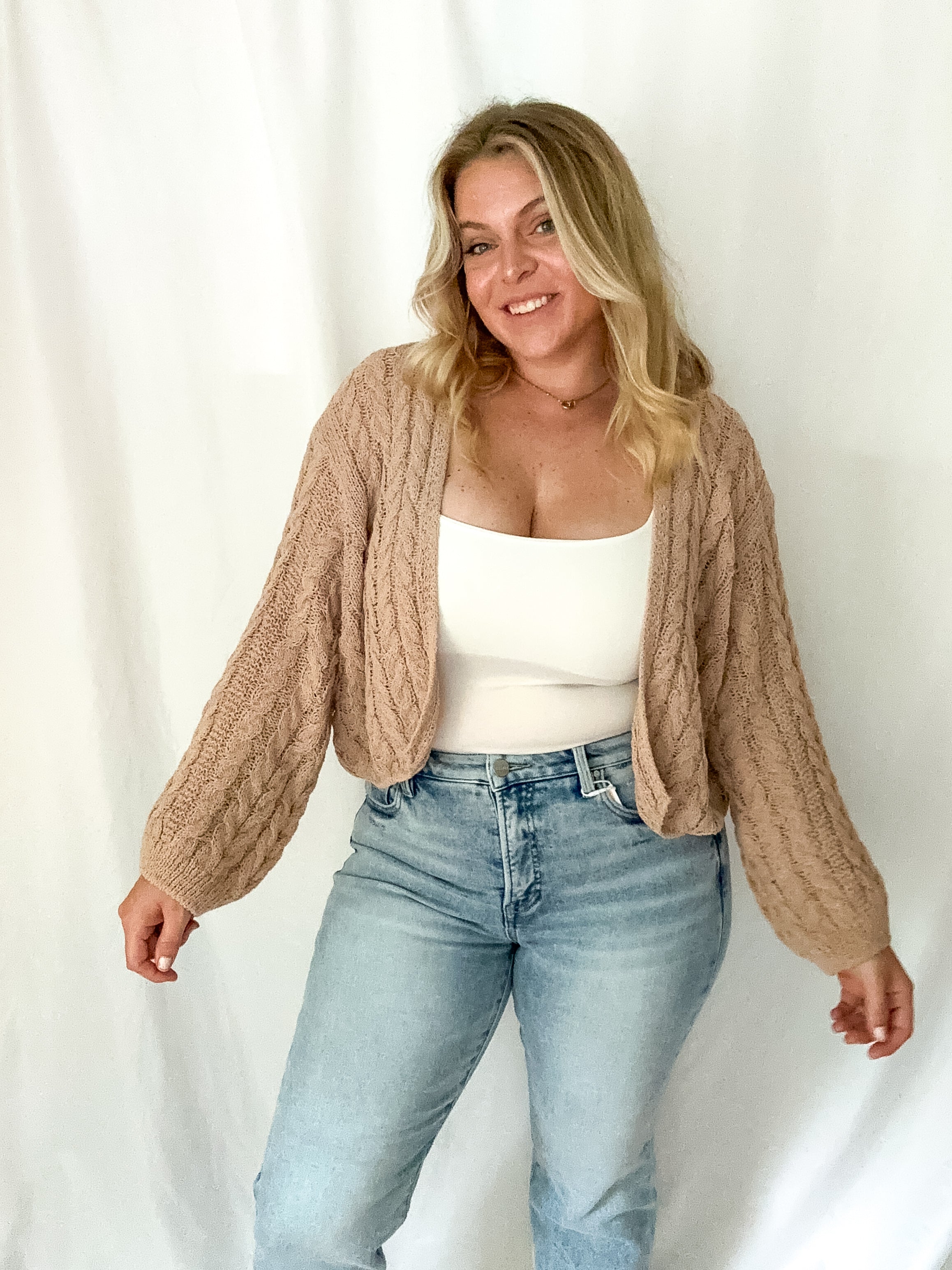Woman wearing a beige cable knit cardigan, white top, and light blue jeans against a white background