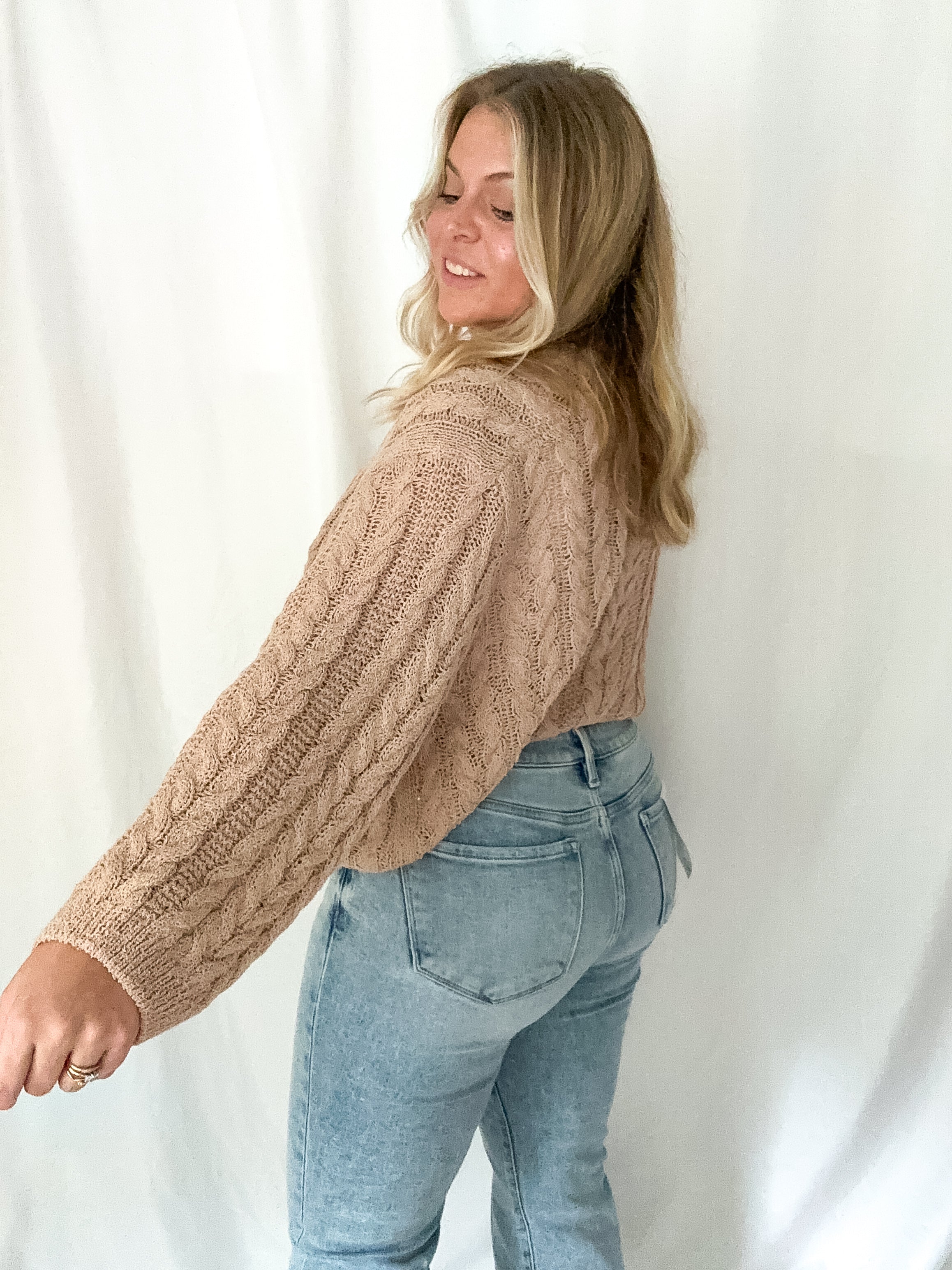 Woman wearing a beige cable knit sweater and blue jeans against a white background