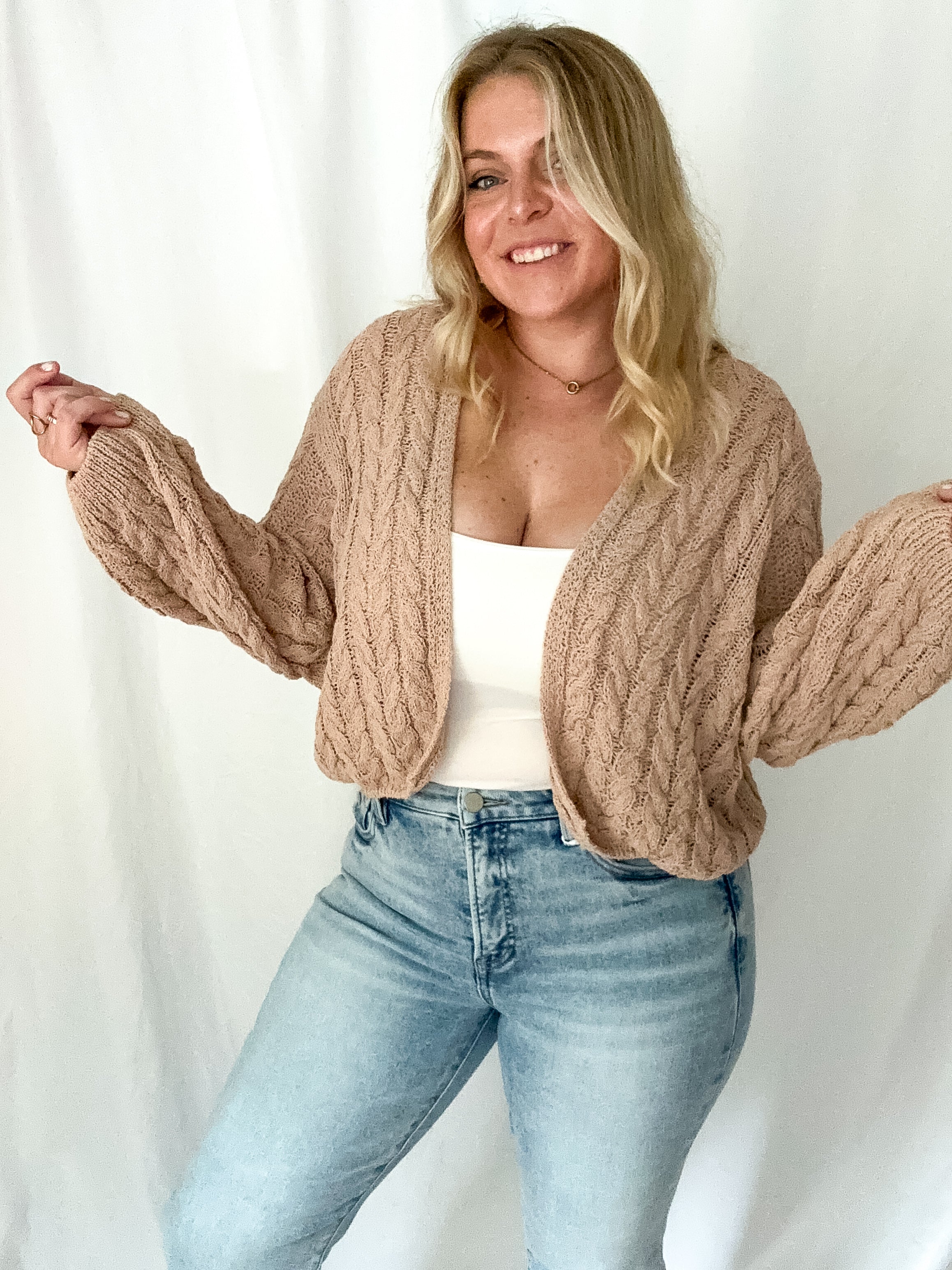 Woman wearing a beige cable knit cardigan over a white top and blue jeans on a white background