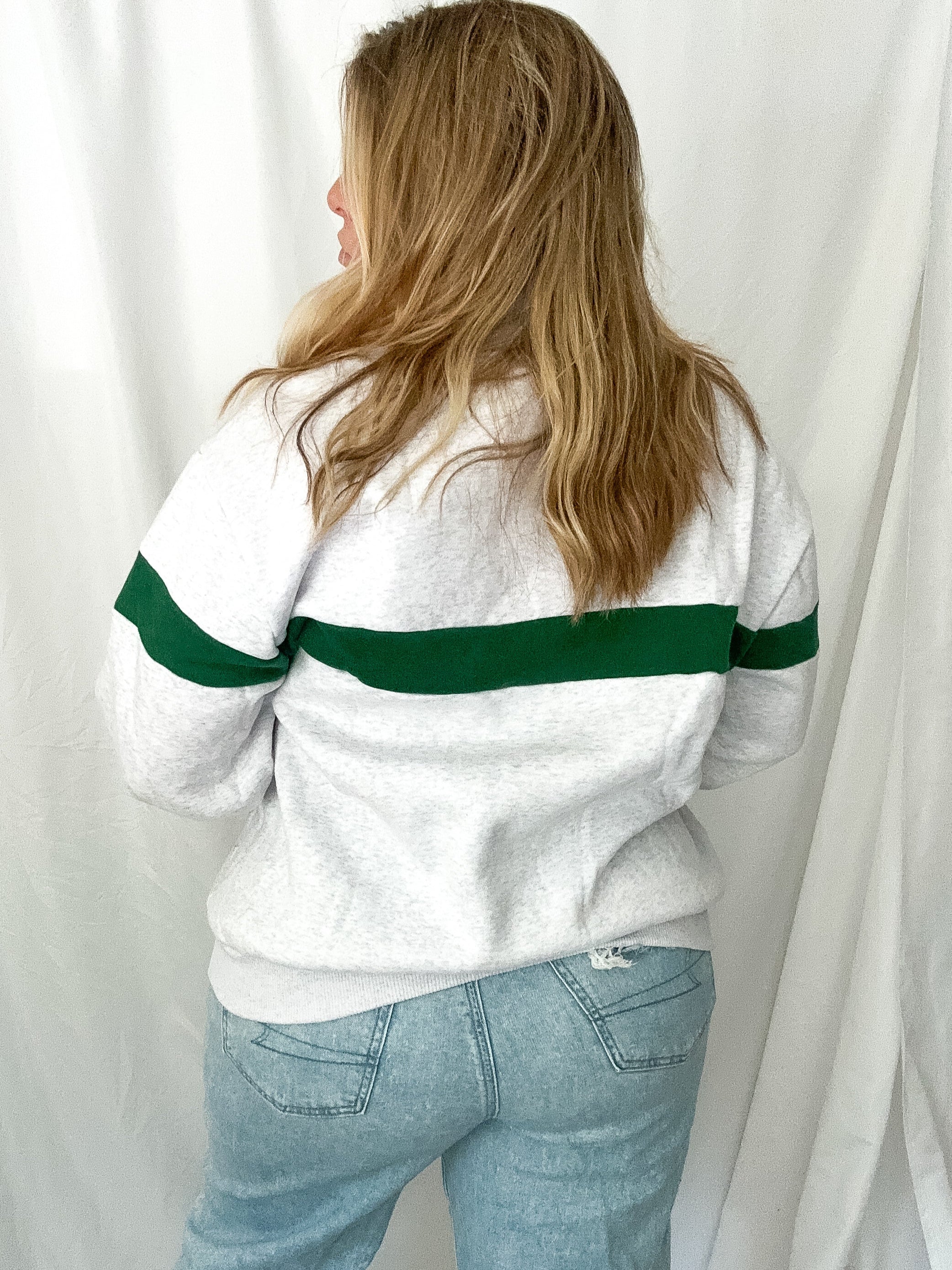 Person wearing a white sweater with green stripes and light blue jeans against a white curtain background