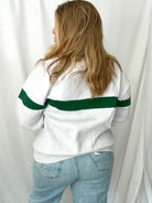 Person wearing a white sweater with green stripes and light blue jeans against a white curtain background