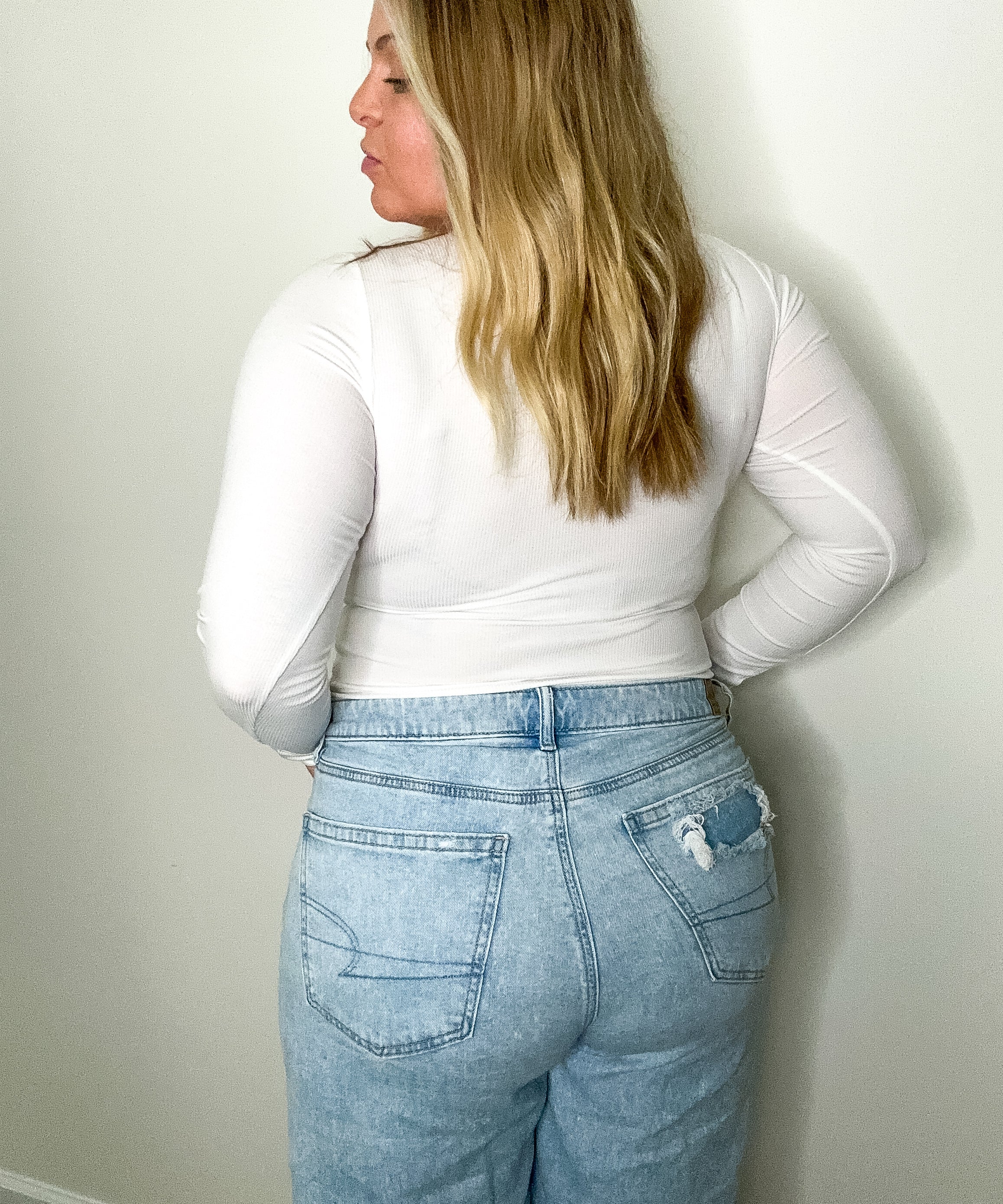 Person wearing a white long-sleeve top and light blue jeans against a plain wall.
