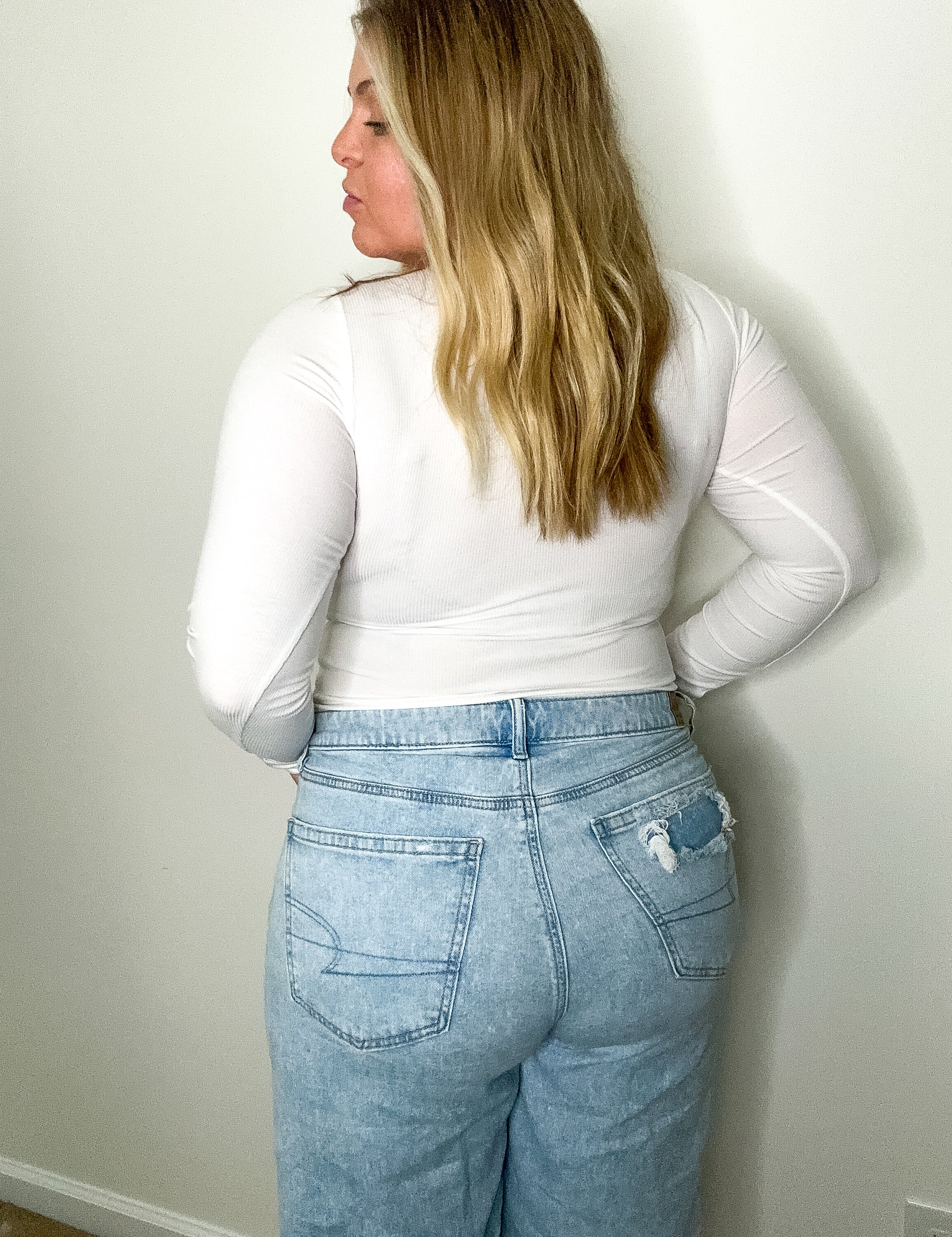 Person wearing a white long-sleeve top and light blue jeans against a plain wall.