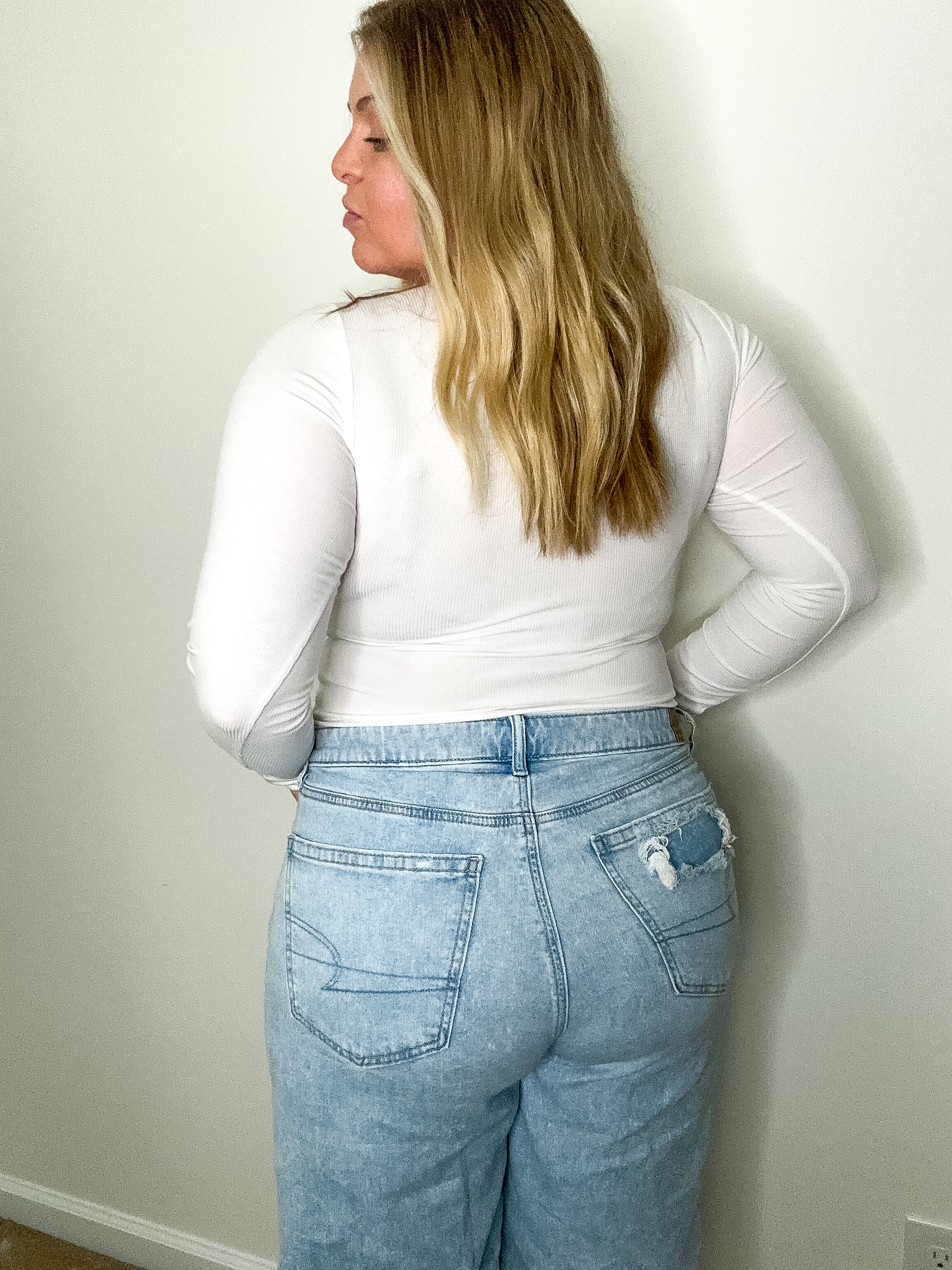 Person wearing a white long-sleeve top and light blue jeans against a plain wall.