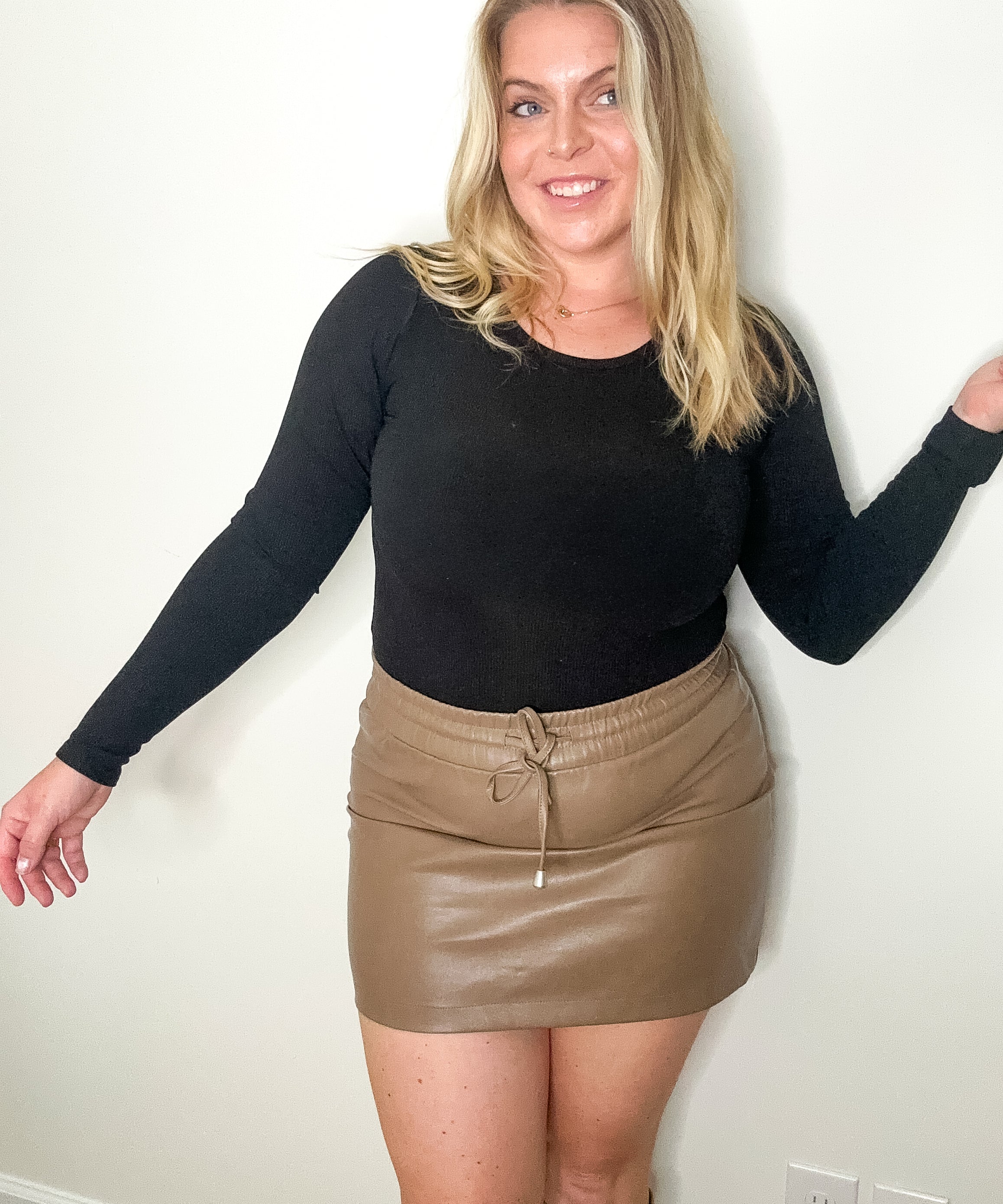 Woman wearing a black long-sleeve top and beige leather skirt against a white wall.