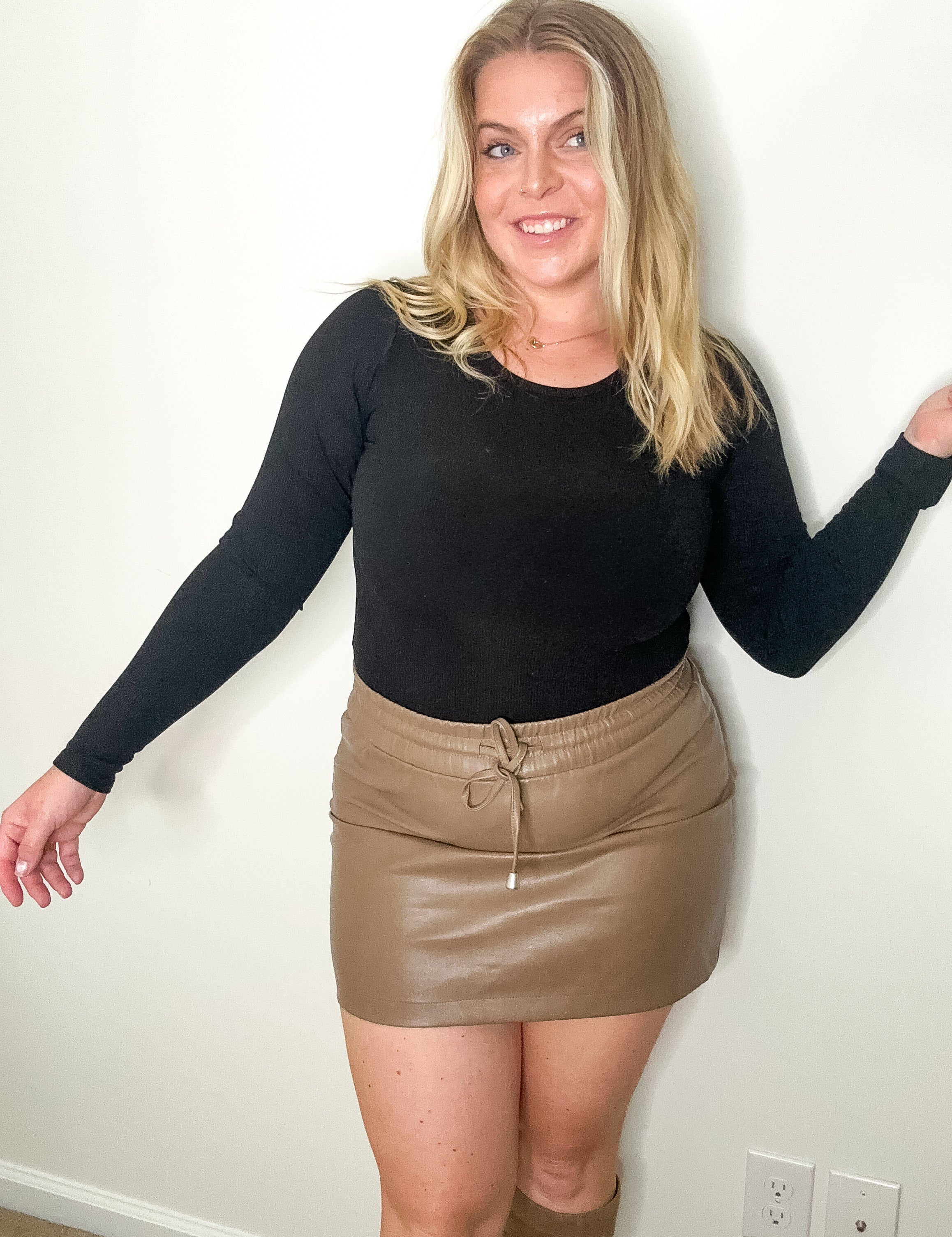 Woman wearing a black long-sleeve top and beige leather skirt against a white wall.