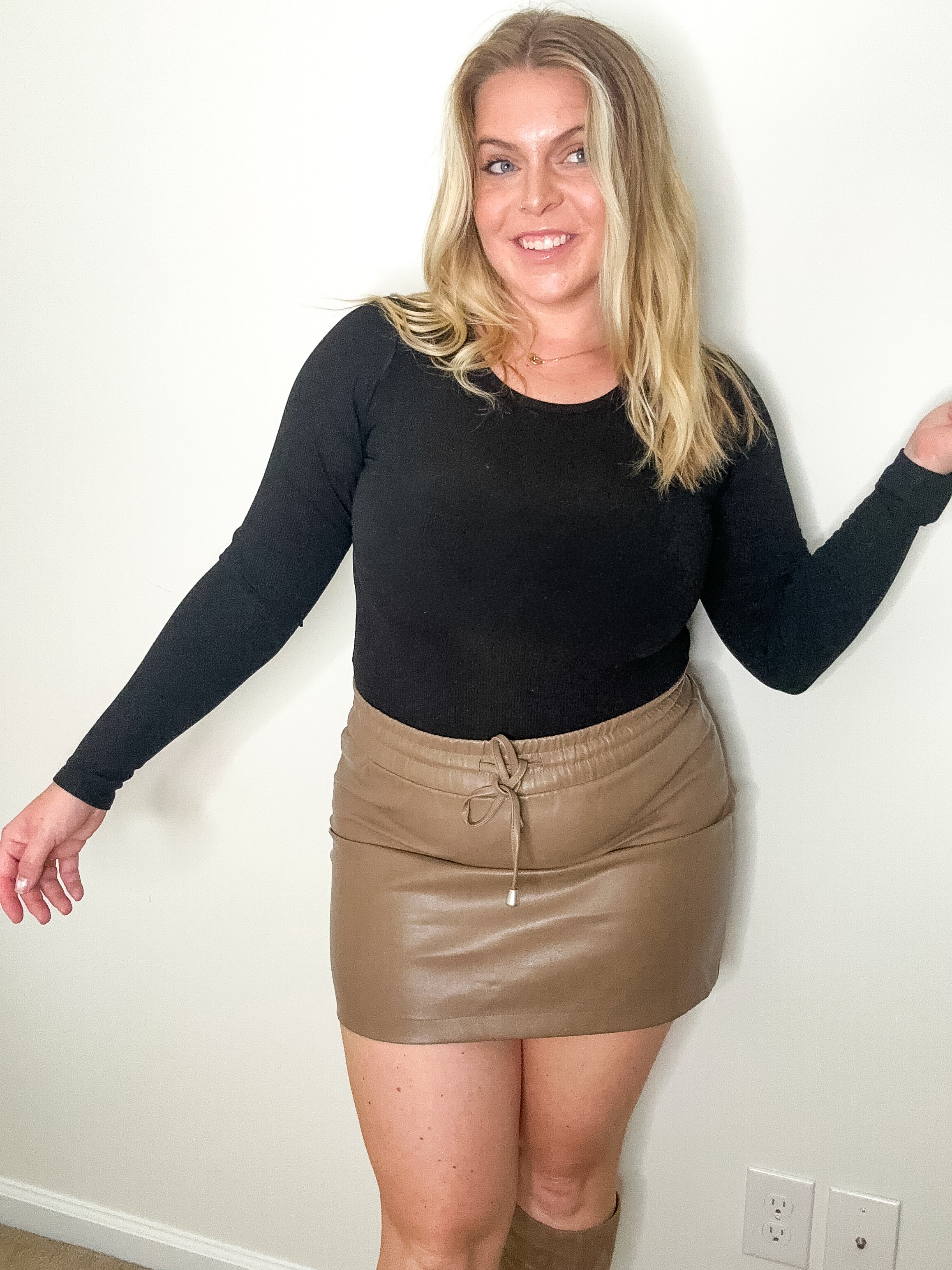 Woman wearing a black long-sleeve top and beige leather skirt against a white wall.