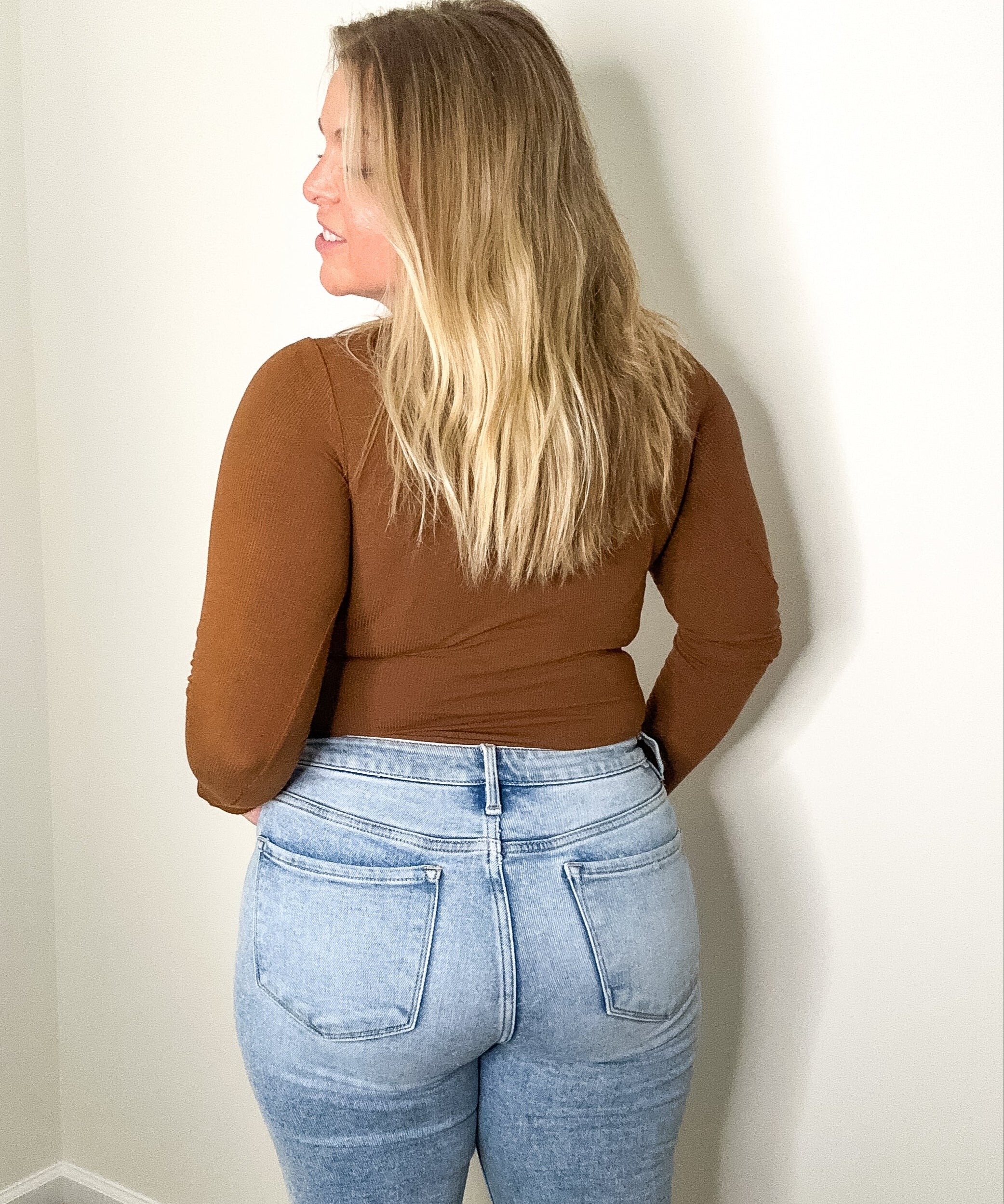 Person wearing a brown top and blue jeans against a plain wall.
