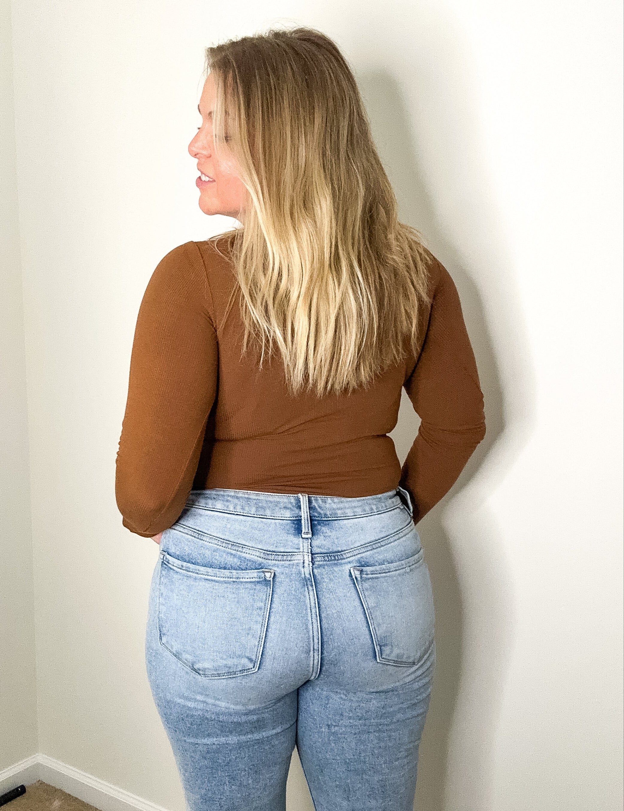 Person wearing a brown top and blue jeans against a plain wall.